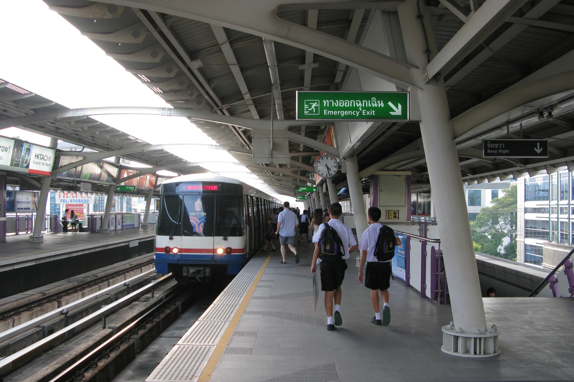 Trem da BTS parado ao lado de plataforma elevada, com passageiros e placas bilíngues em Bangkok.