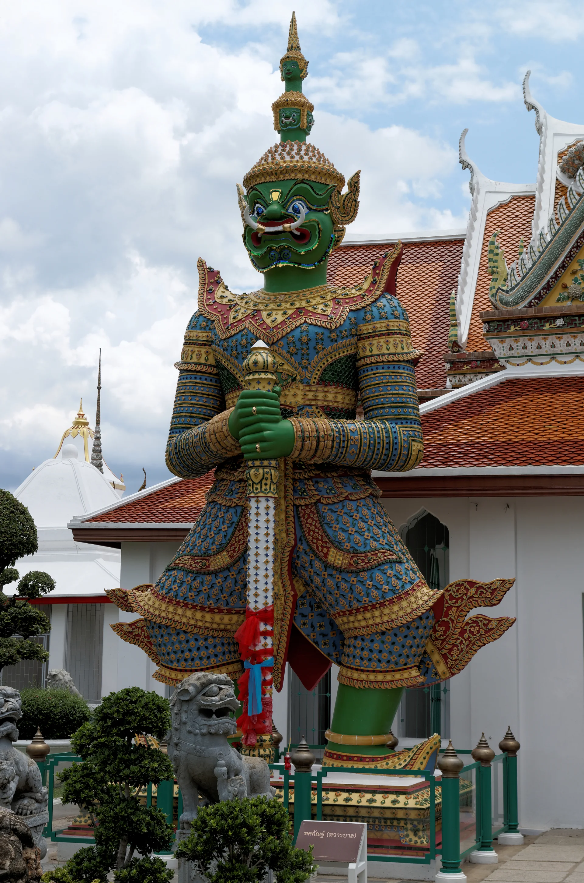 Estátua colorida de Thotsakan, guardião do Wat Arun, diante de telhado com telhas vermelhas e céu azul.