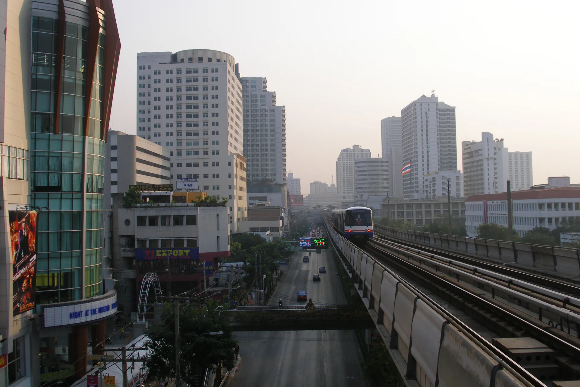 Trem da BTS em via elevada sobre uma avenida, cercado por edifícios do centro de Bangkok ao fim da tarde.
