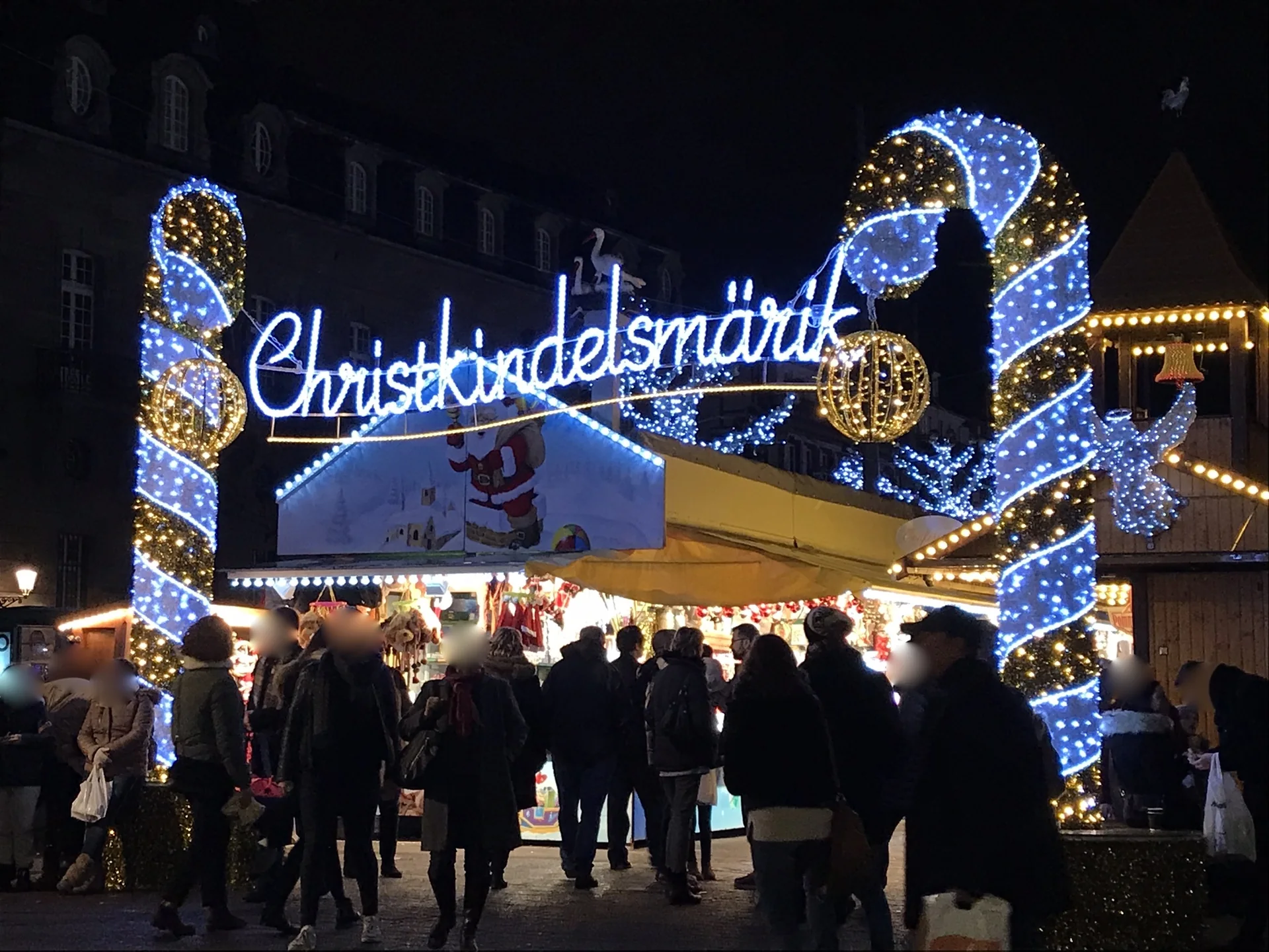 Entrada iluminada do mercado de Natal de Estrasburgo à noite, com visitantes sob a decoração Chrìstkìndelsmärik.