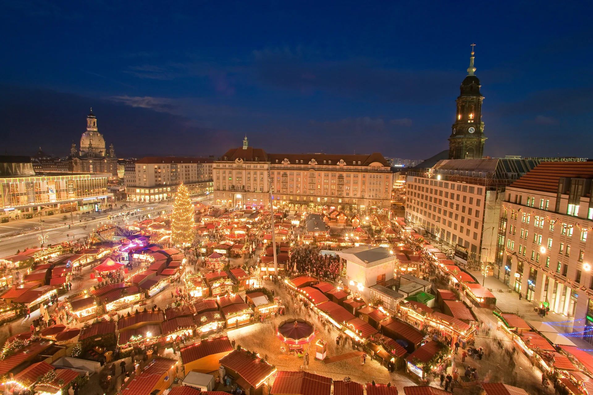 Vista elevada da Altmarkt de Dresden iluminada durante o Striezelmarkt, com chalés, árvore de Natal e a silhueta da cidade ao anoitecer.