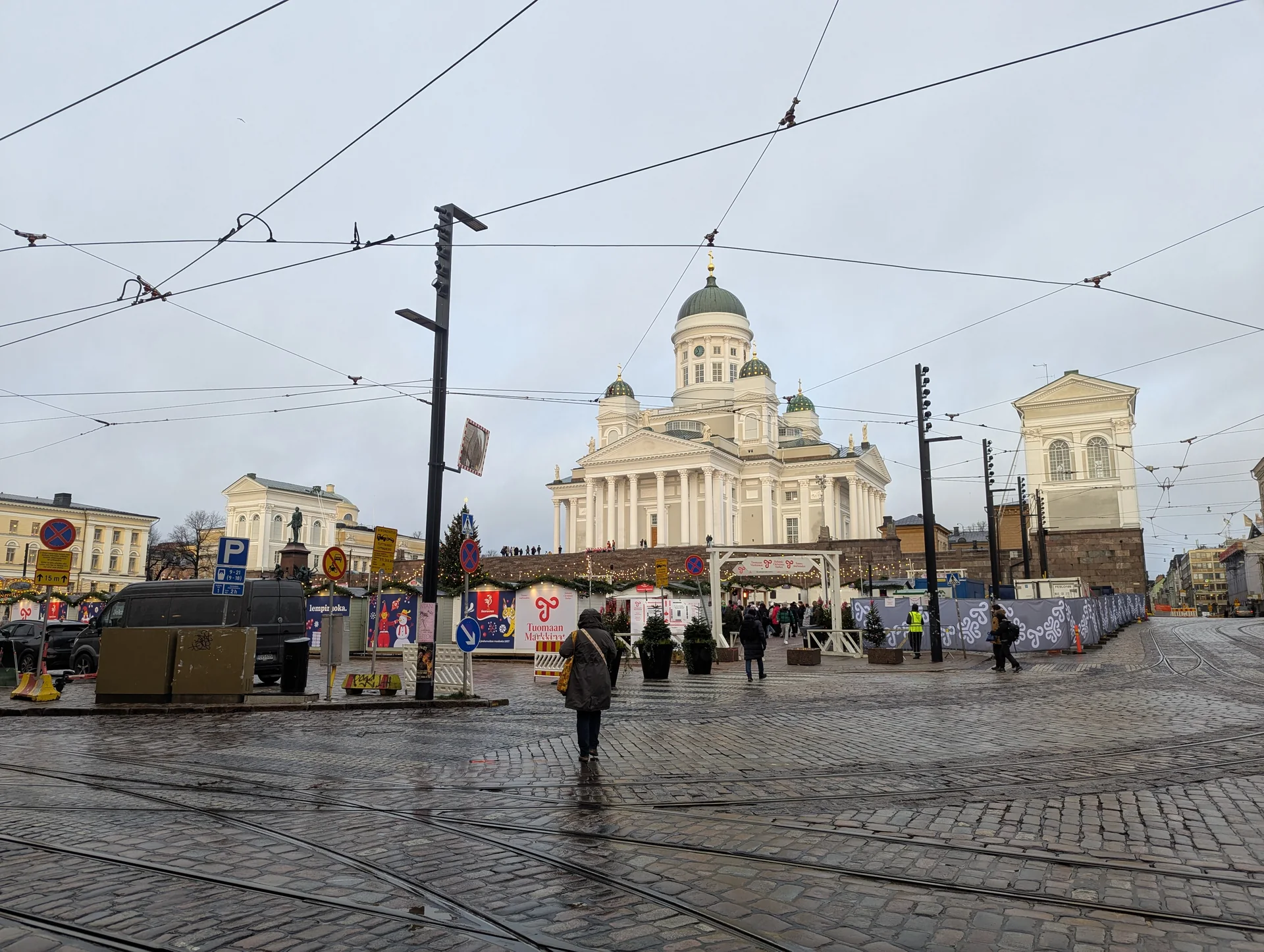 Praça do Senado de Helsinque em dia cinzento, com a Catedral de Helsinque acima da entrada do Mercado de Natal e trilhos de bonde no primeiro plano.