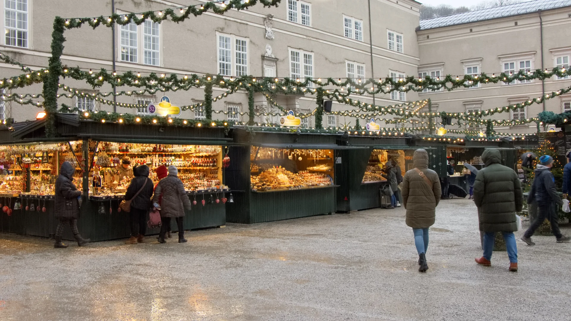 Bancas iluminadas e visitantes sob chuva leve no Christkindlmarkt da Domplatz, no centro histórico de Salzburgo.