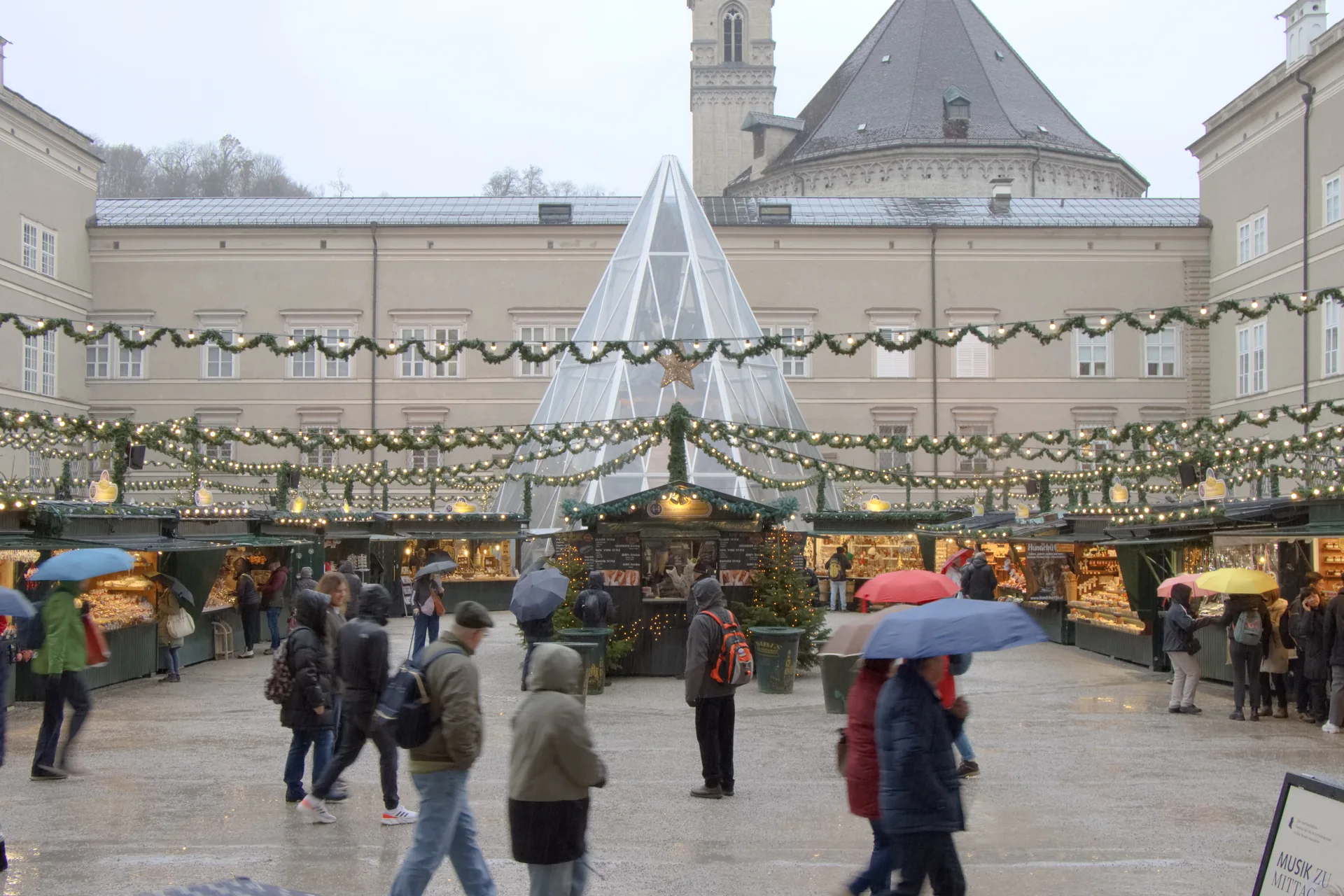 Vista da Domplatz com as bancas do Christkindlmarkt, árvore iluminada e visitantes com guarda-chuvas em Salzburgo.