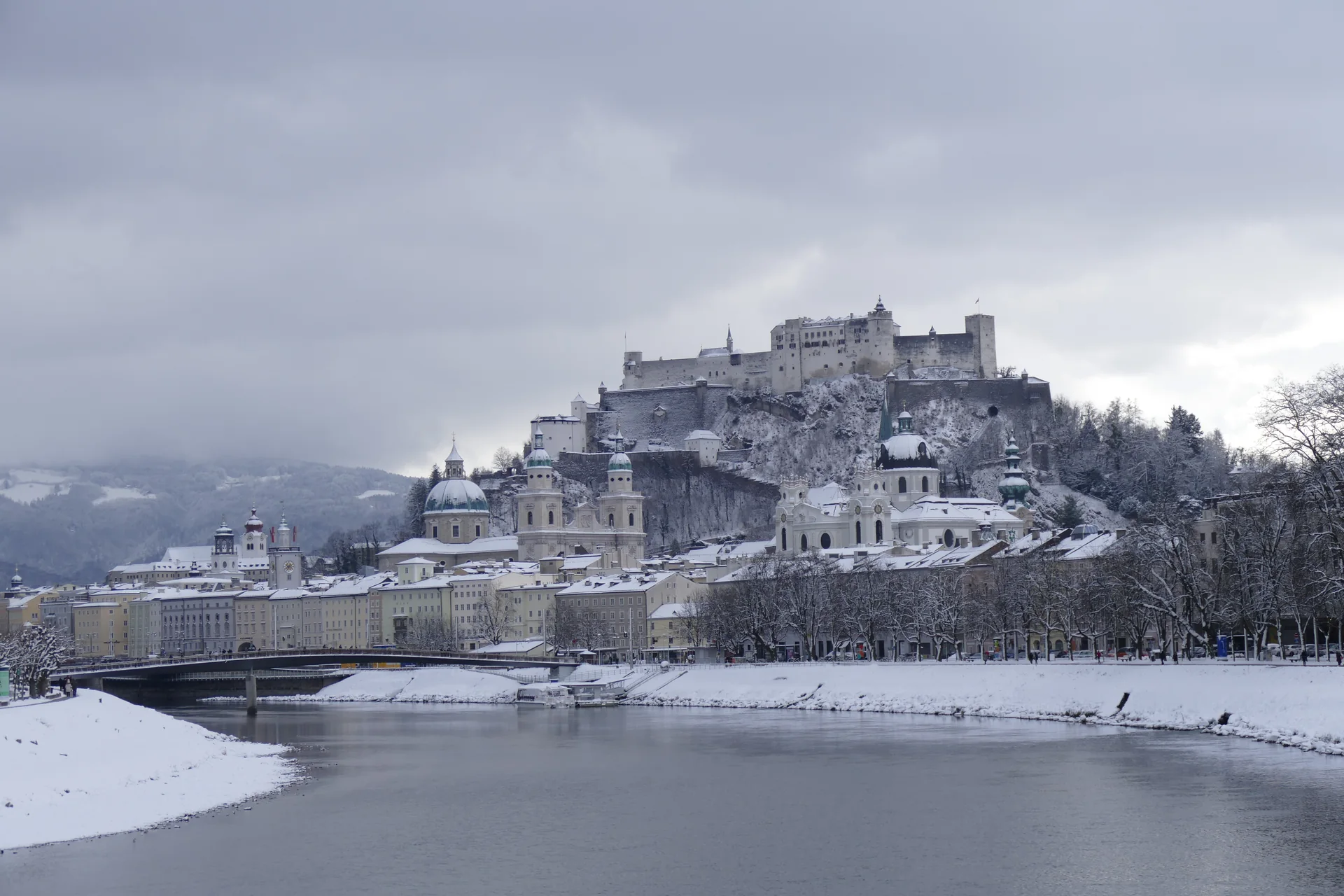 Centro histórico de Salzburgo coberto de neve, com o rio Salzach, torres e a Fortaleza Hohensalzburg ao fundo.