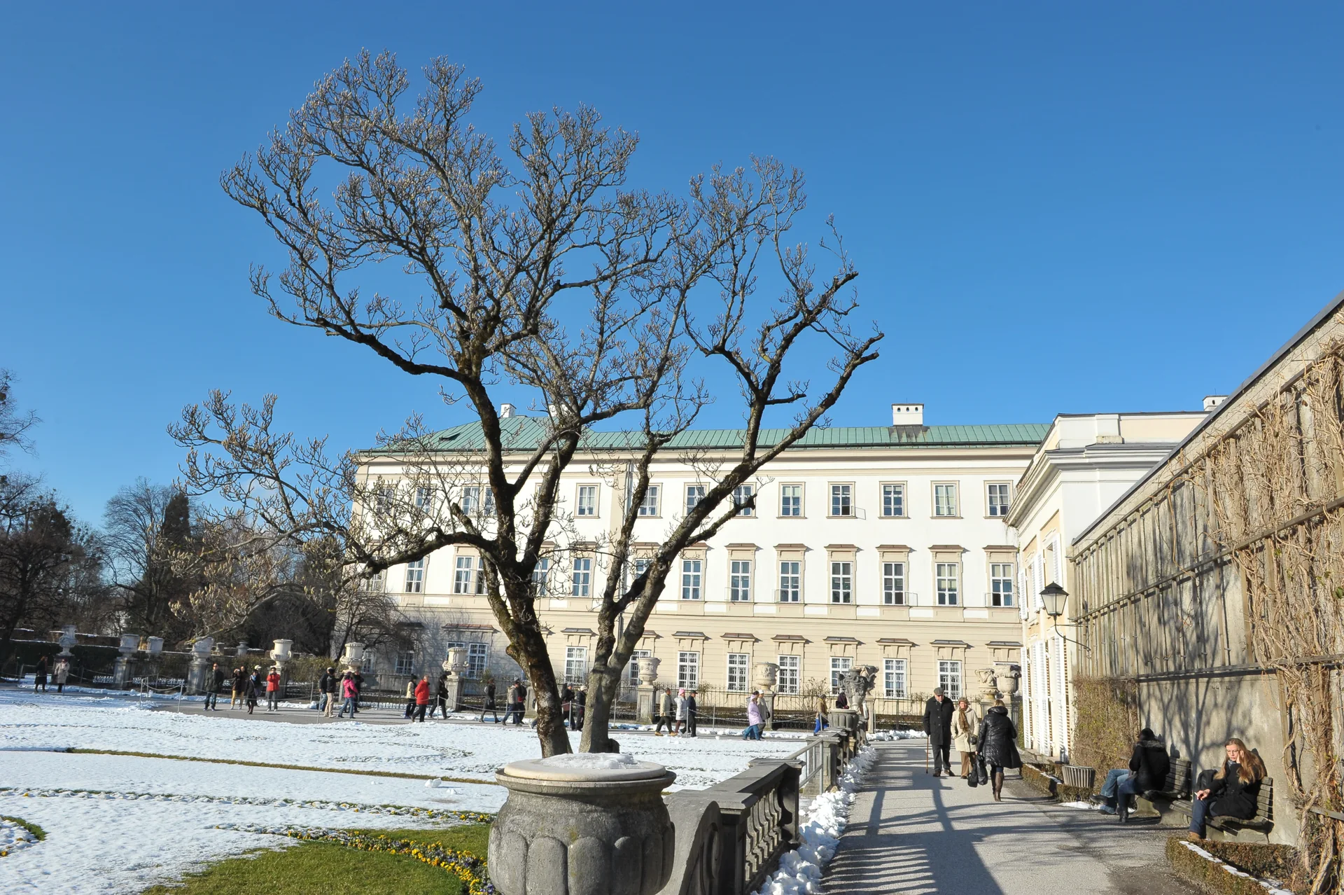 Jardim de Mirabell com neve, visitantes e a fachada do Palácio de Mirabell sob céu azul em Salzburgo.
