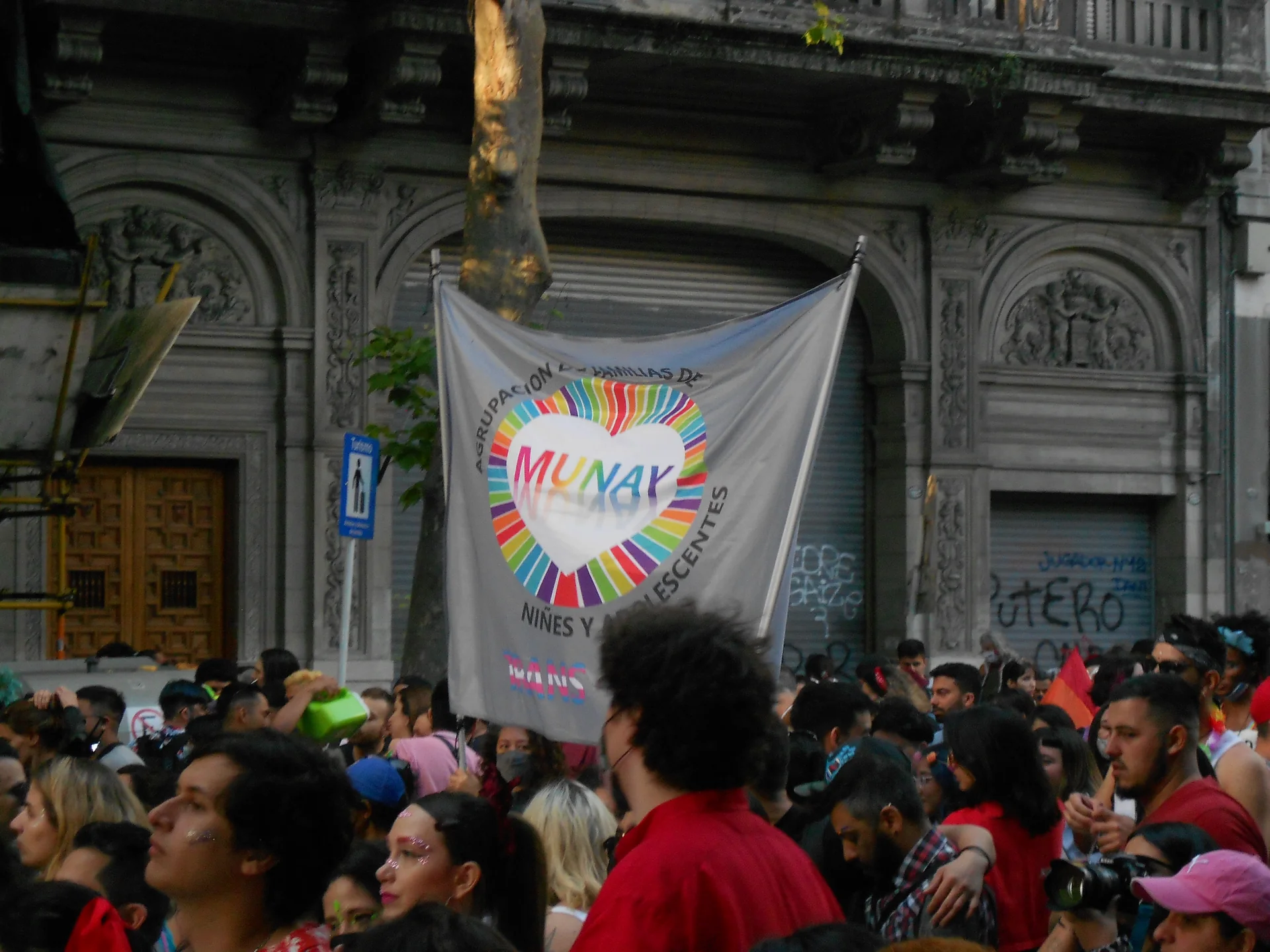 Pessoas reunidas na XXX Marcha del Orgulho LGBTIQ+ de Buenos Aires, diante de edifício histórico, com faixa de coletivo comunitário