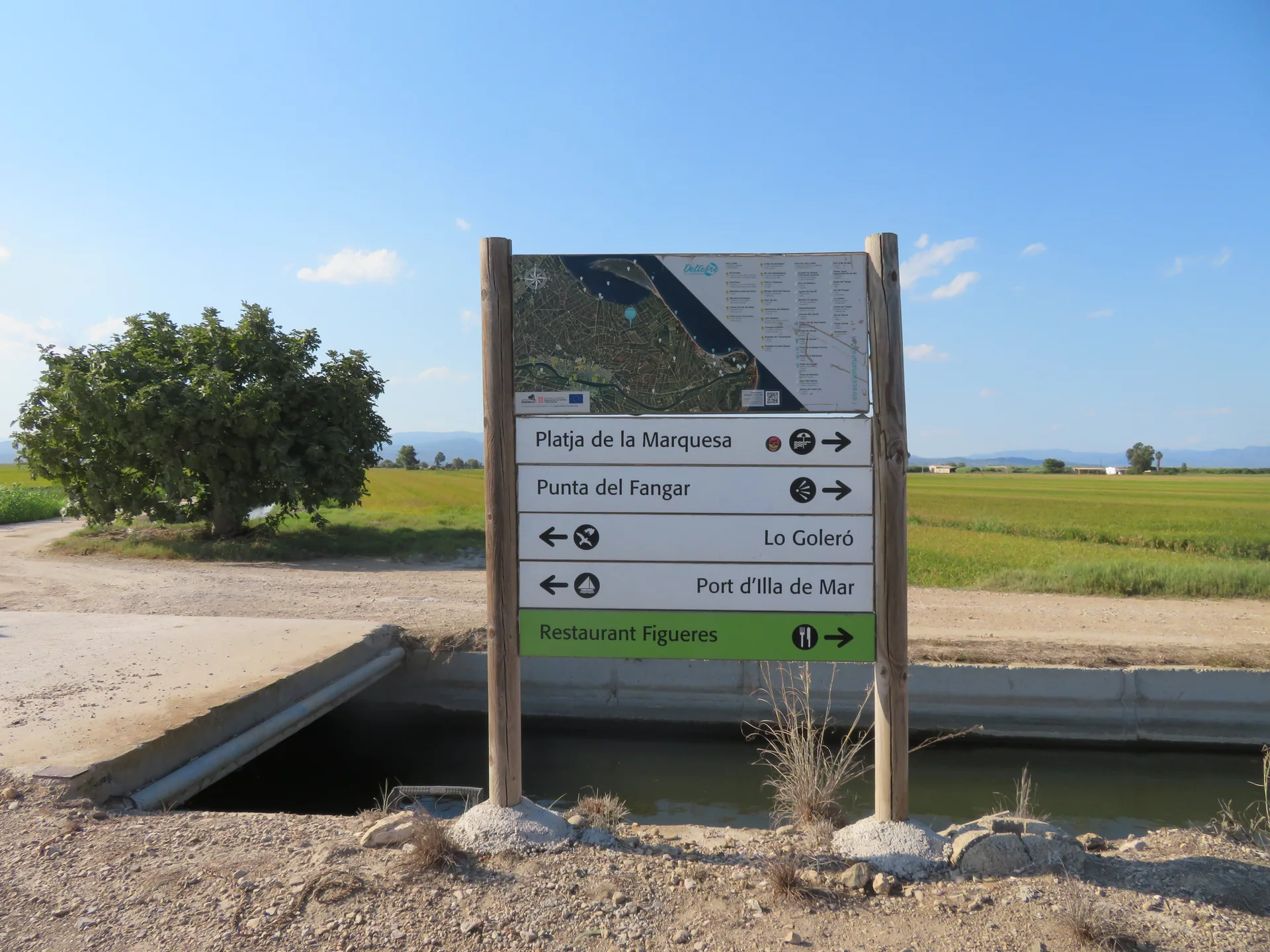 Placa de orientação junto a um canal e campos no Delta do Ebro, sob céu claro.