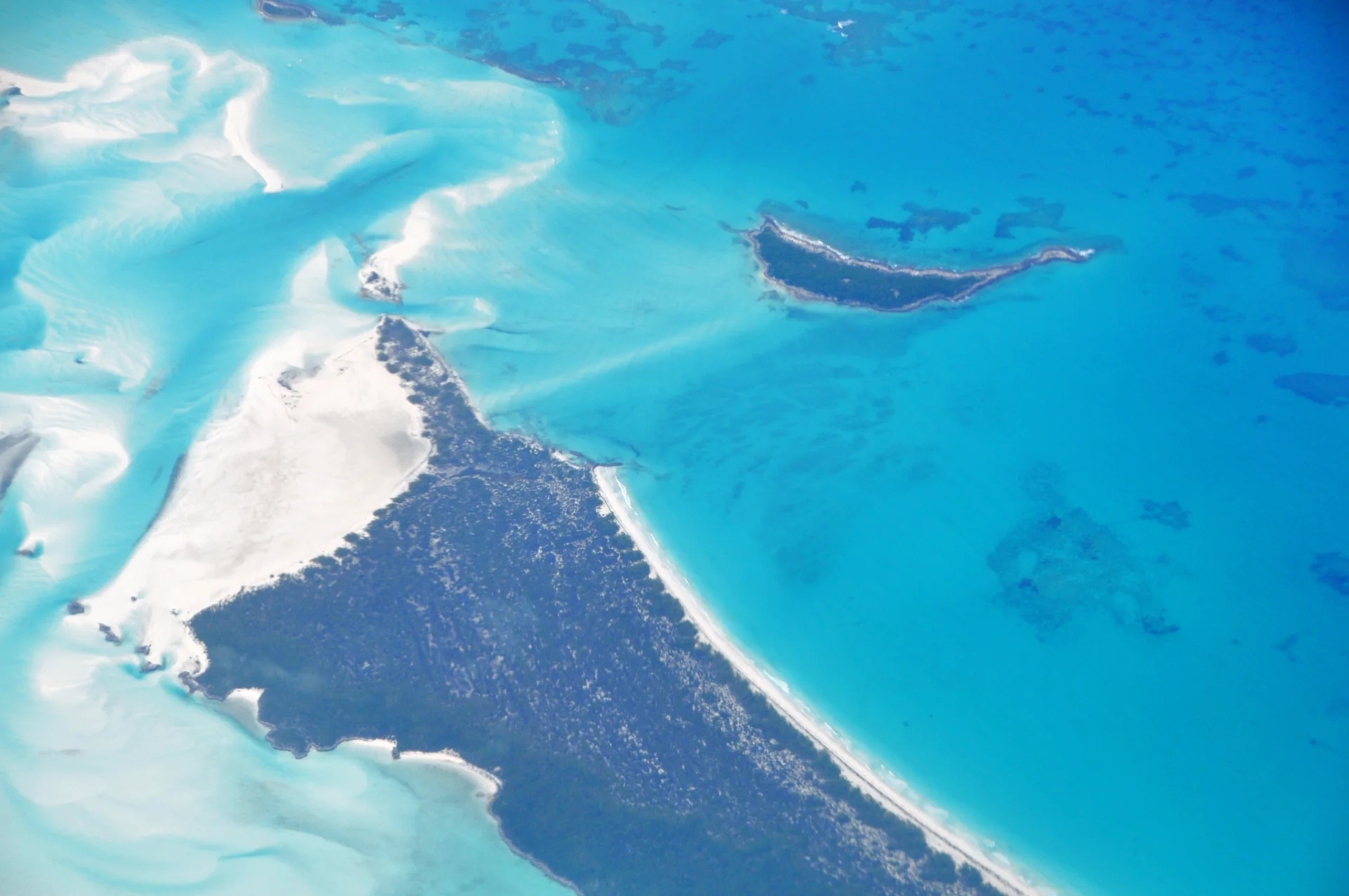 Vista aérea ensolarada de Hawksbill Cay, com areia branca, vegetação baixa e água azul-turquesa nas Exumas.