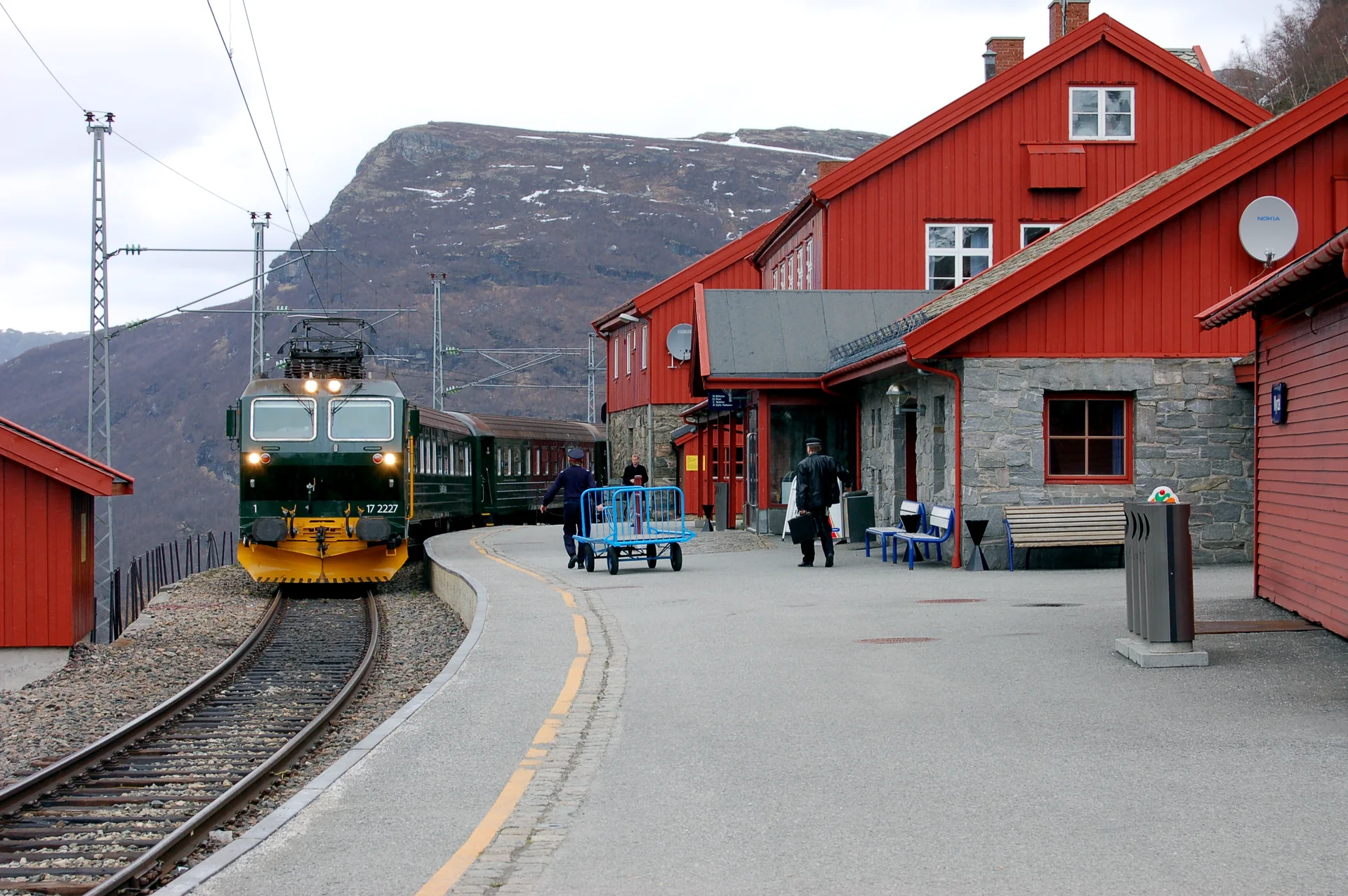 Trem da Flåmsbana estacionado em Myrdal, ao lado da plataforma e de um edifício vermelho sob montanhas.