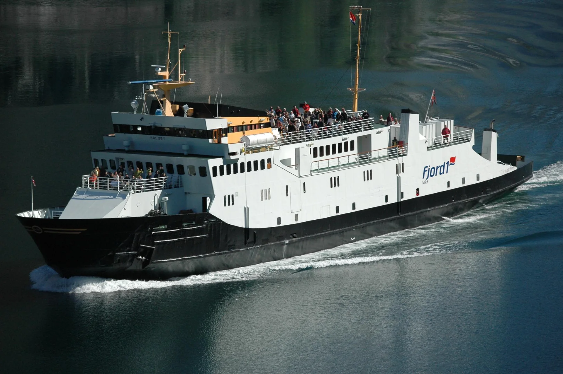 Ferry MF Bolsøy navega nas águas escuras do Geirangerfjord sob luz natural.