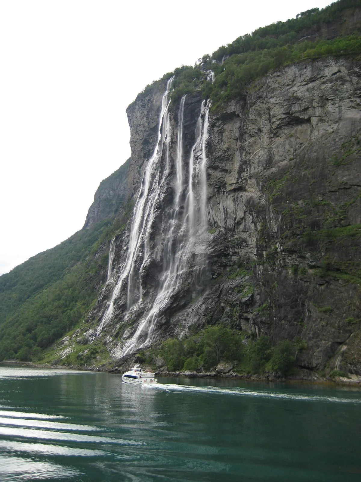 Cachoeiras das Sete Irmãs descem pela encosta rochosa até o Geirangerfjord, com barco navegando abaixo.