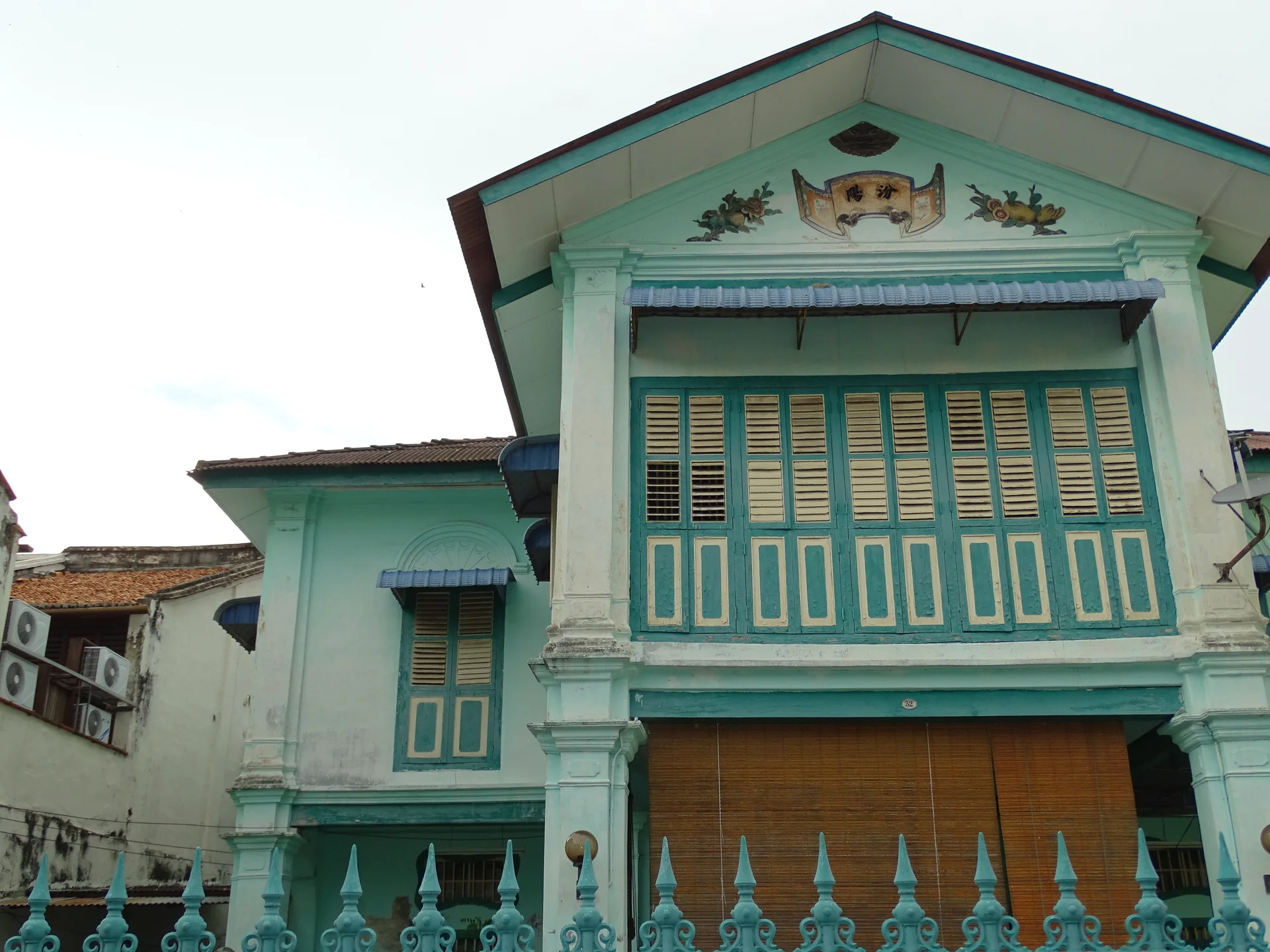 Detalhe de uma casa histórica verde-água em George Town, com venezianas, frontão e gradil sob luz diurna.