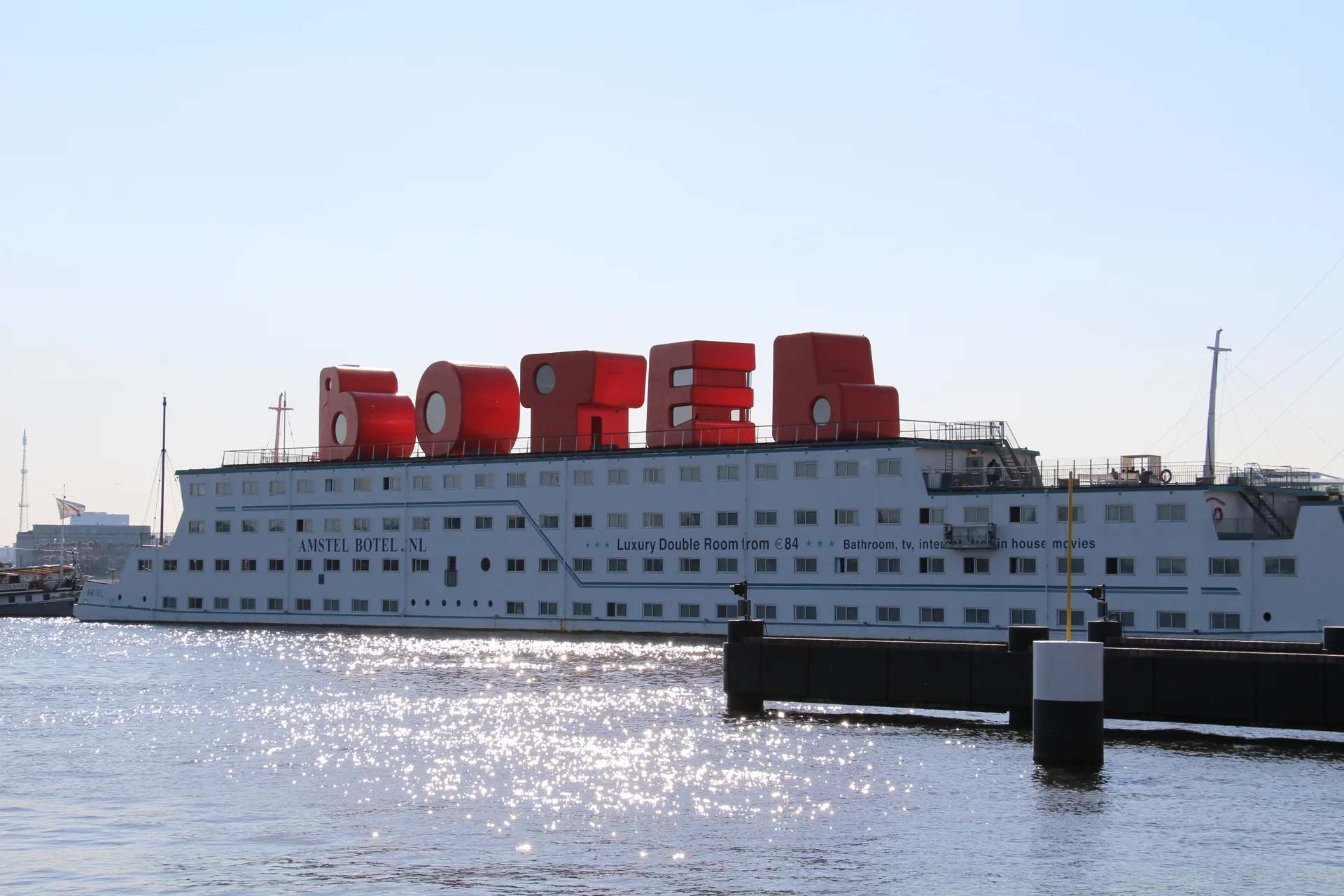 Navio branco do Botel atracado no IJ, com letras vermelhas no convés e água brilhando ao sol.