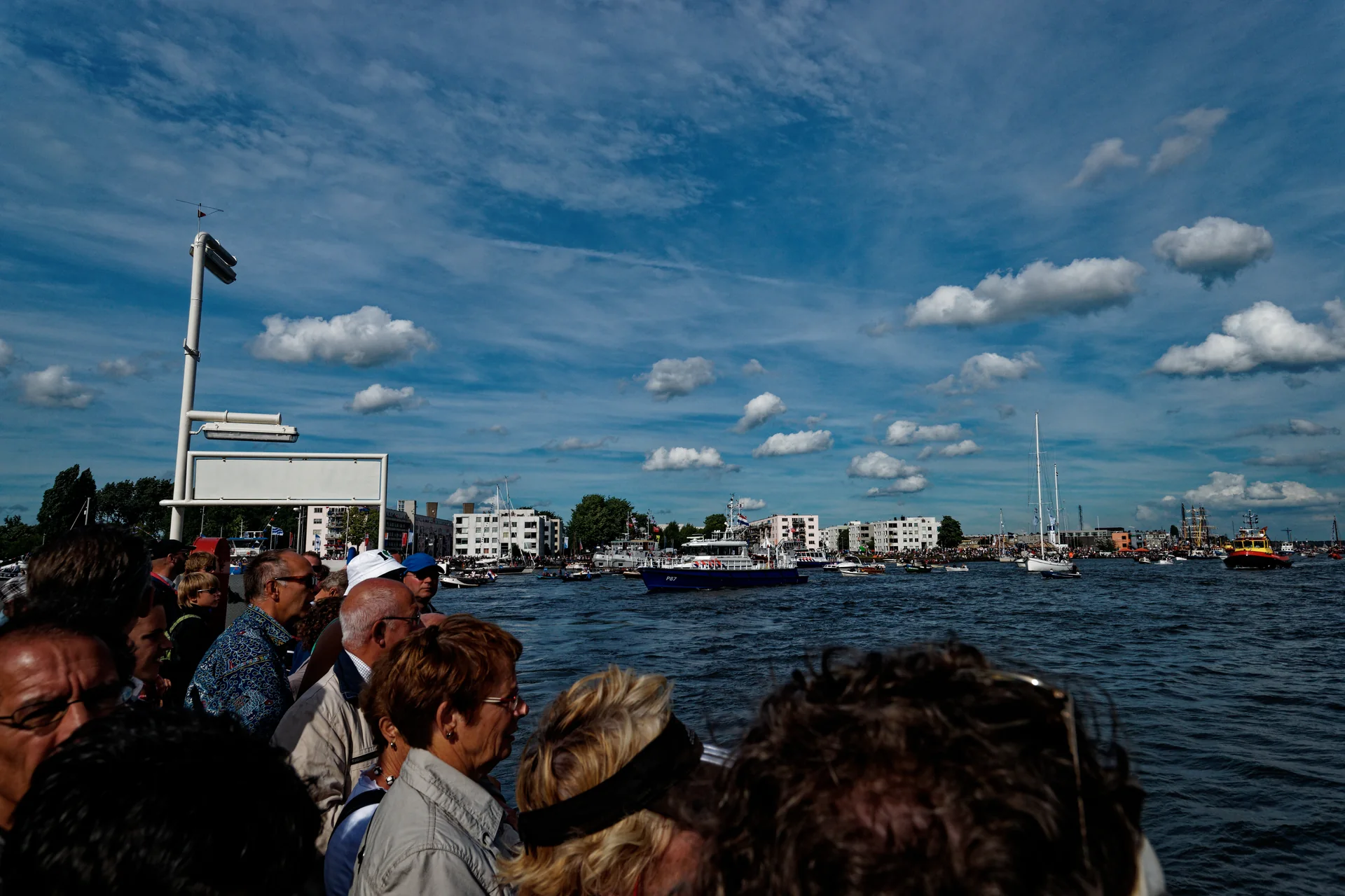 Passageiros reunidos em uma balsa no IJ, com céu azul e embarcações no evento Sail Amsterdam.