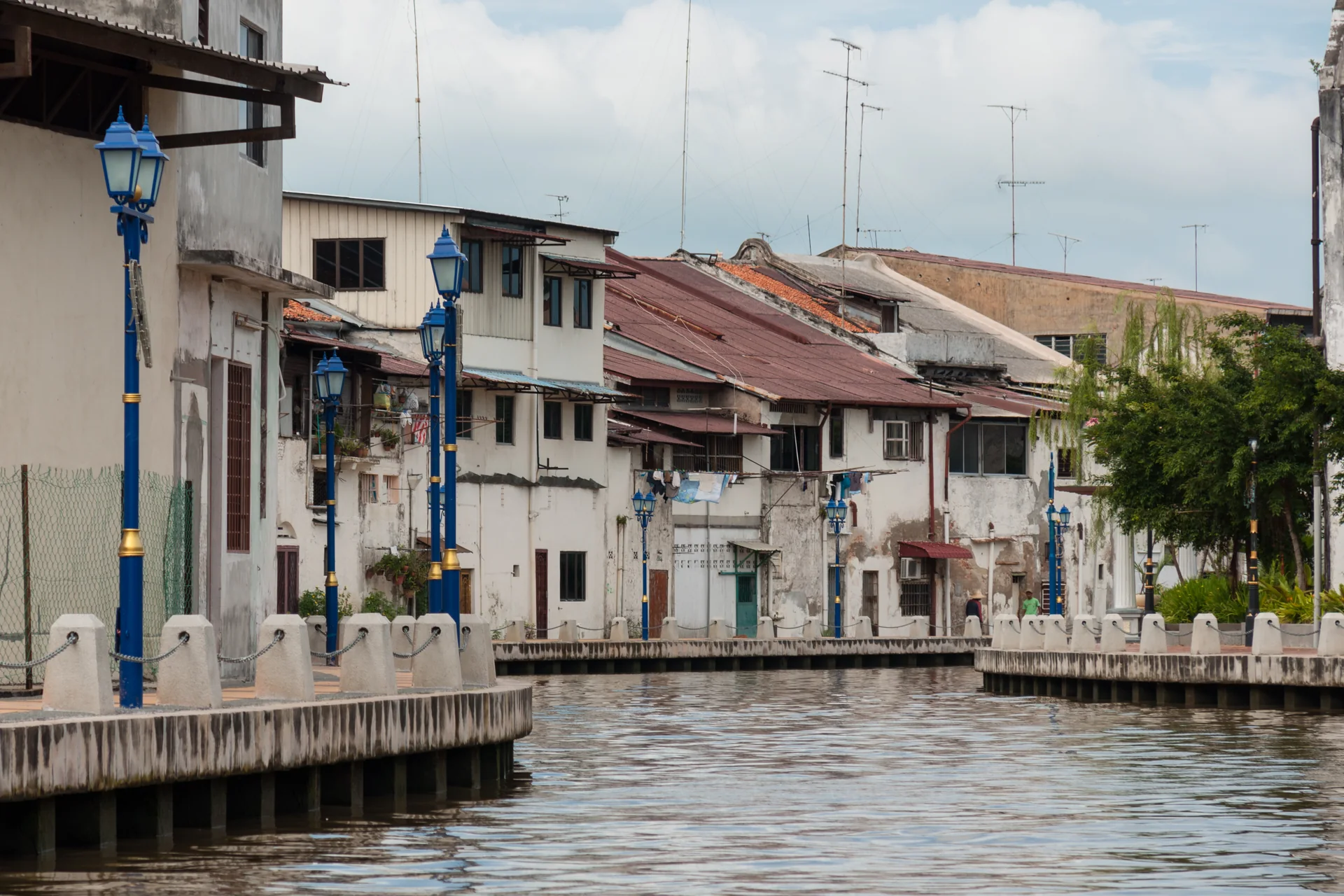 Casas históricas e luminárias azuis junto à margem do Rio Melaka durante o dia.