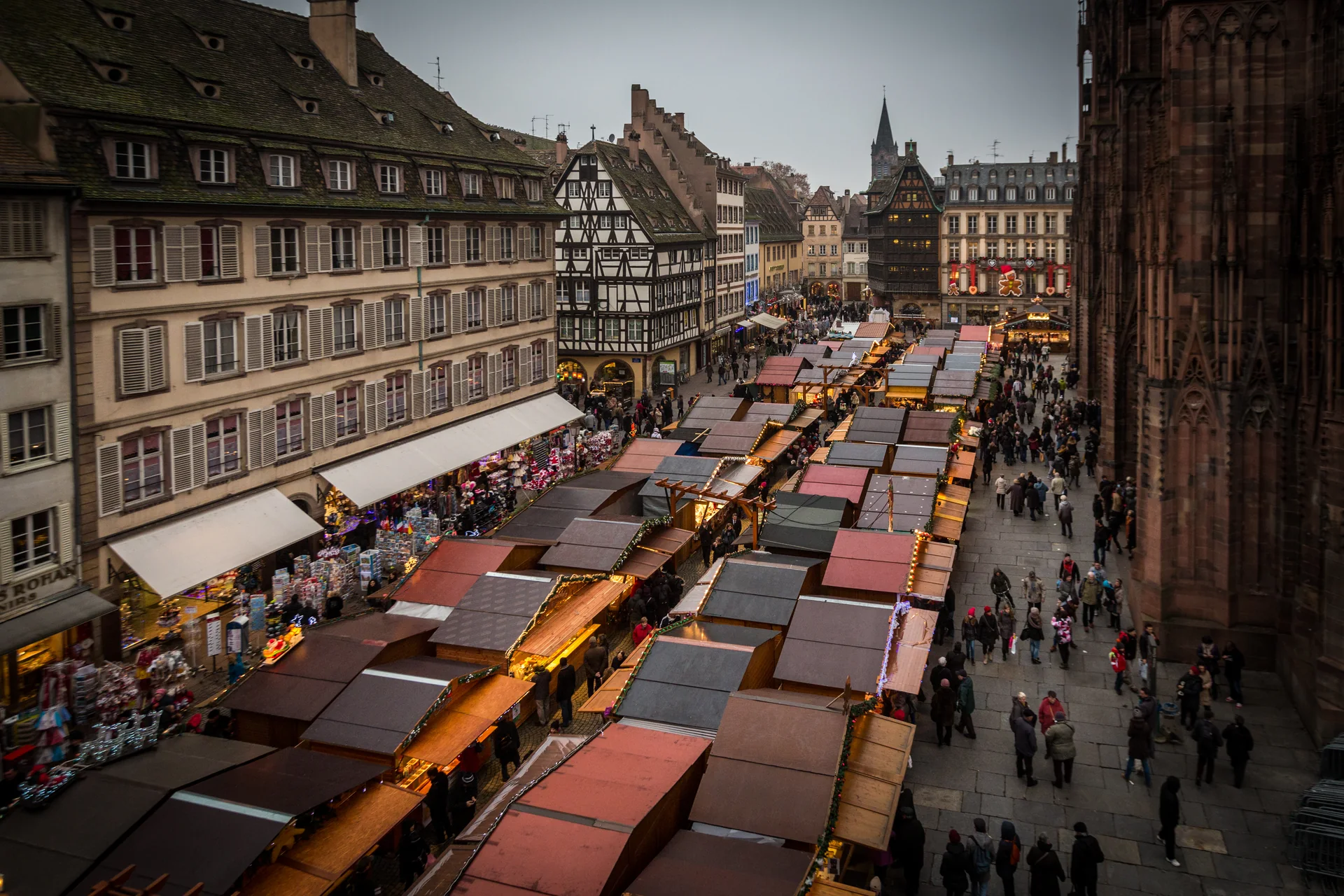 Vista elevada dos chalés e visitantes do mercado de Natal junto à Catedral de Estrasburgo, no fim da tarde.