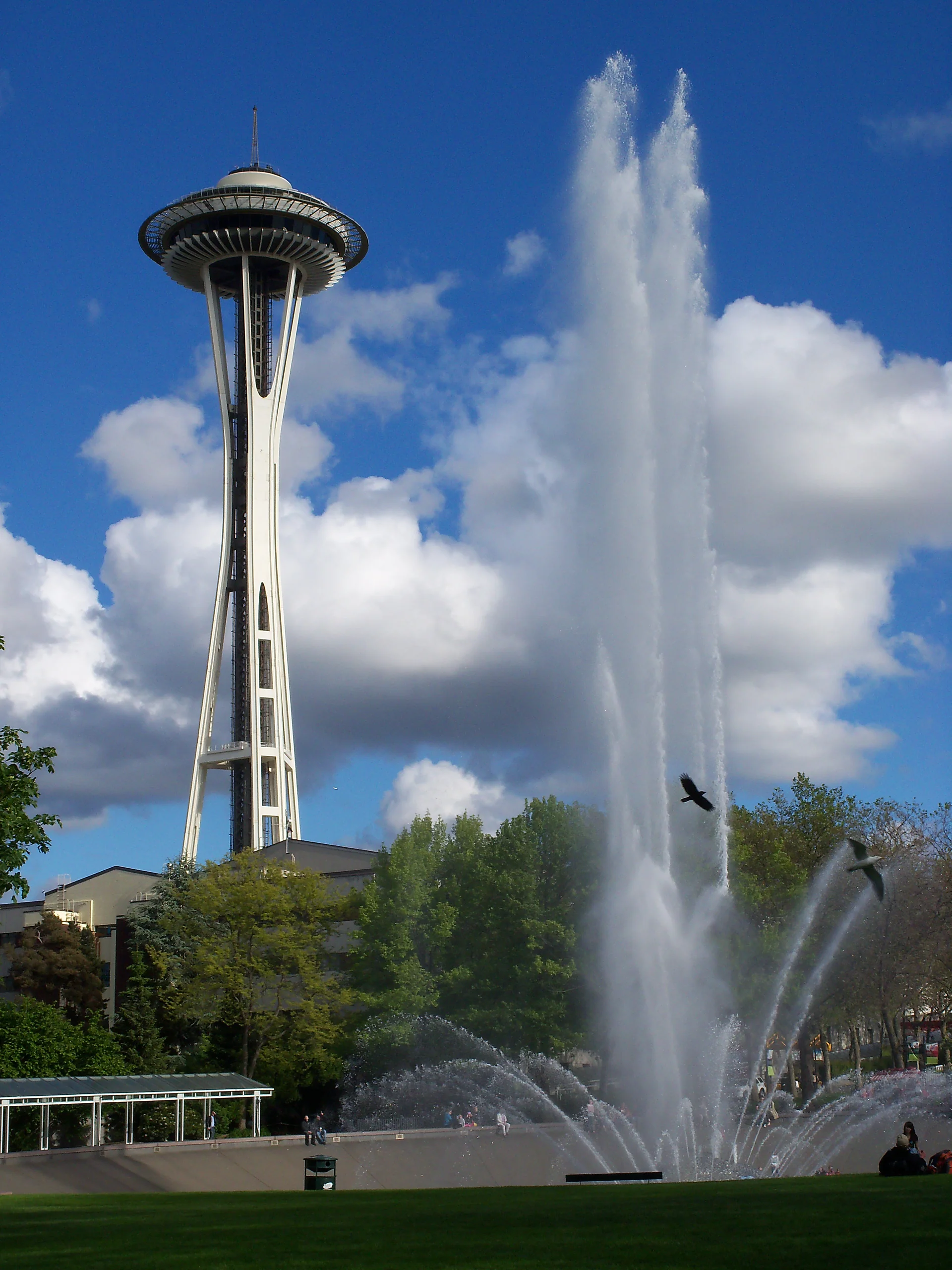 Space Needle e International Fountain em funcionamento no Seattle Center, sob céu azul durante o dia.