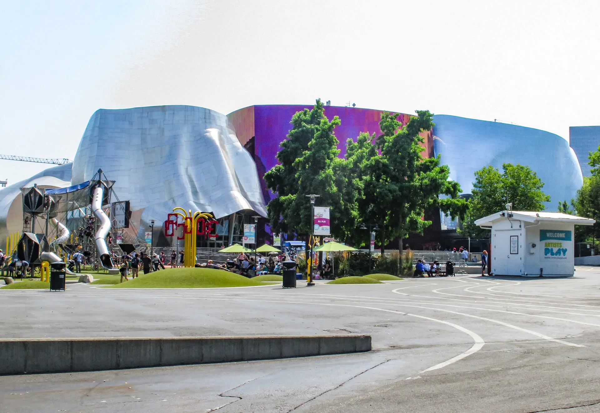 Fachada metálica colorida do Museum of Pop Culture, com visitantes e área verde do Seattle Center durante o dia.