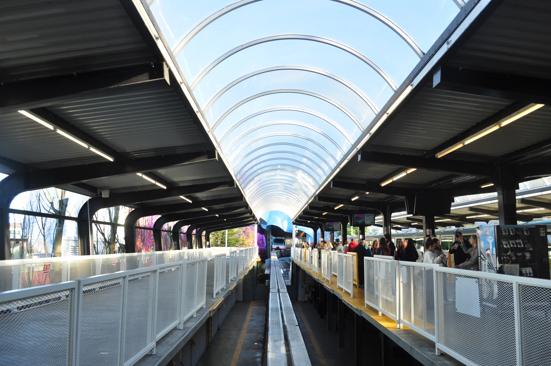 Plataforma do Seattle Center Monorail com passageiros e o MoPOP ao fundo em luz natural.