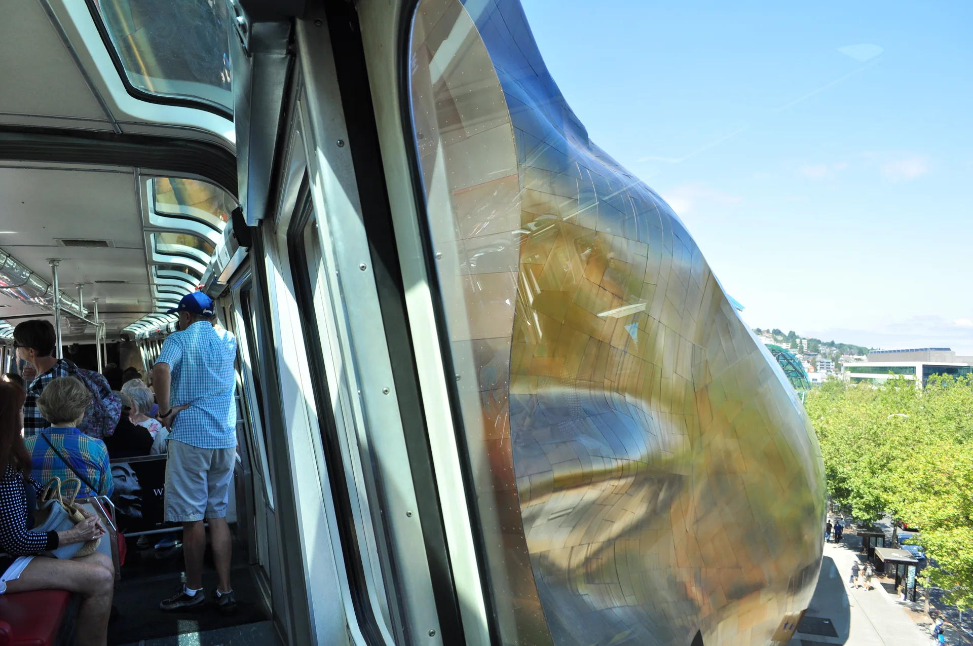 Interior do Seattle Center Monorail com passageiros enquanto o trem passa junto à fachada metálica do MoPOP.