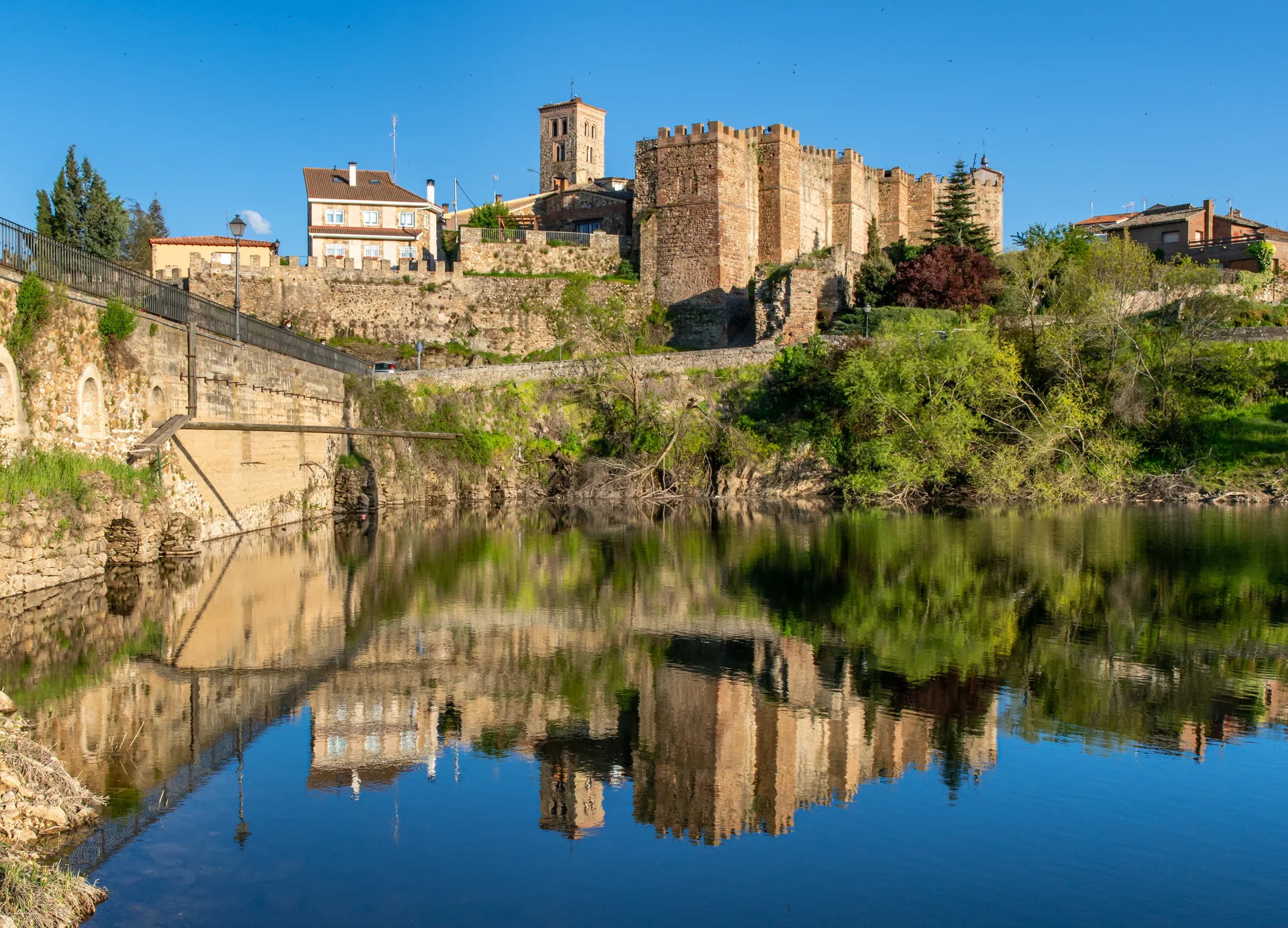 Centro histórico murado de Buitrago del Lozoya refletido no rio Lozoya, visto de uma margem arborizada.