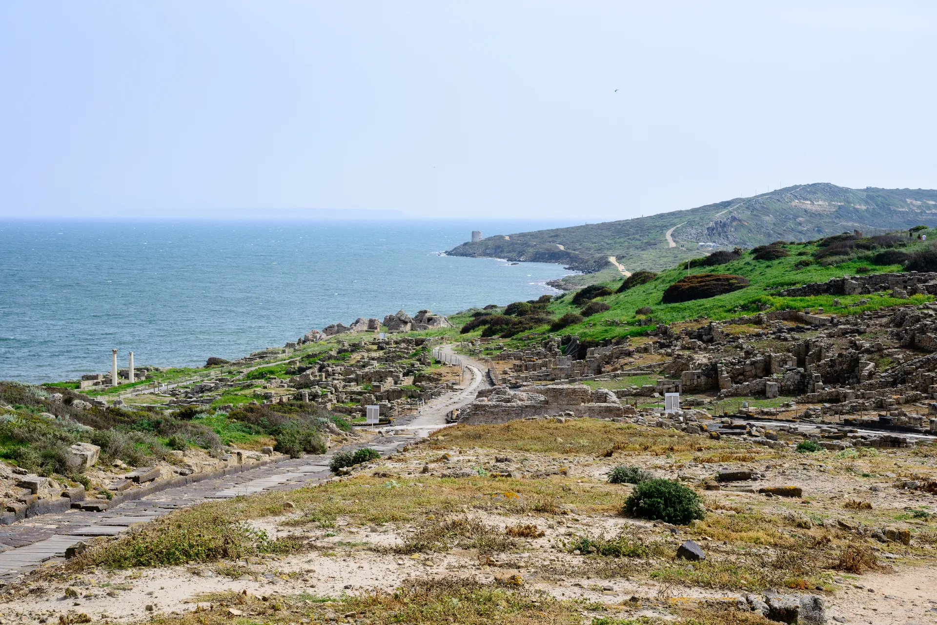 Ruínas arqueológicas de Tharros sobre a costa azul do Sinis, no sul da Sardenha, com colinas ao fundo.