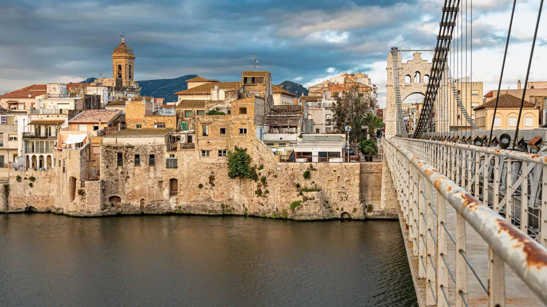 Rio Ebro e ponte suspensa de Amposta vistos ao amanhecer, com o casario histórico da cidade ao fundo.
