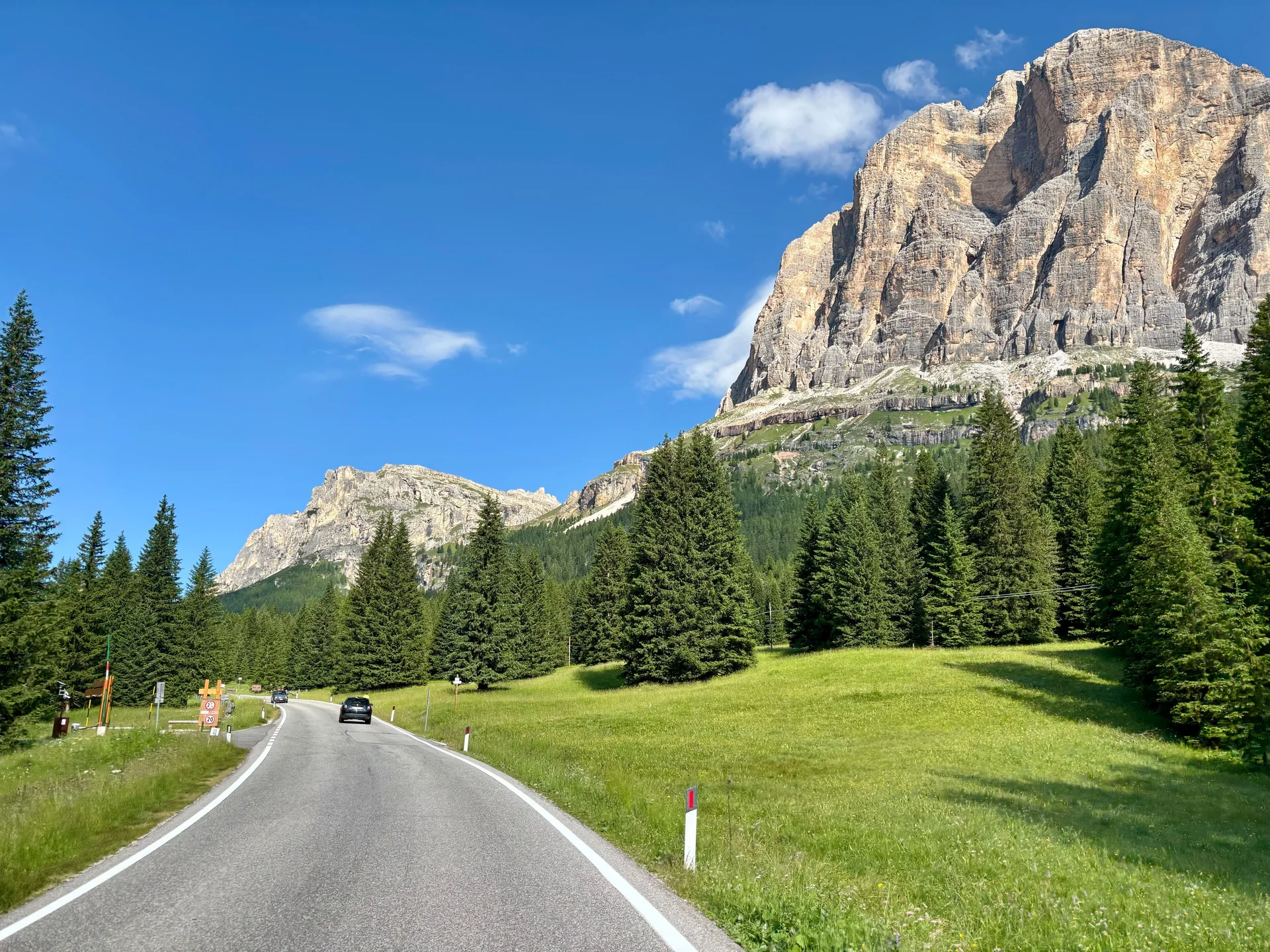 Estrada SR48 em direção ao Passo Falzarego, entre coníferas, prado e paredão dolomítico sob céu azul.