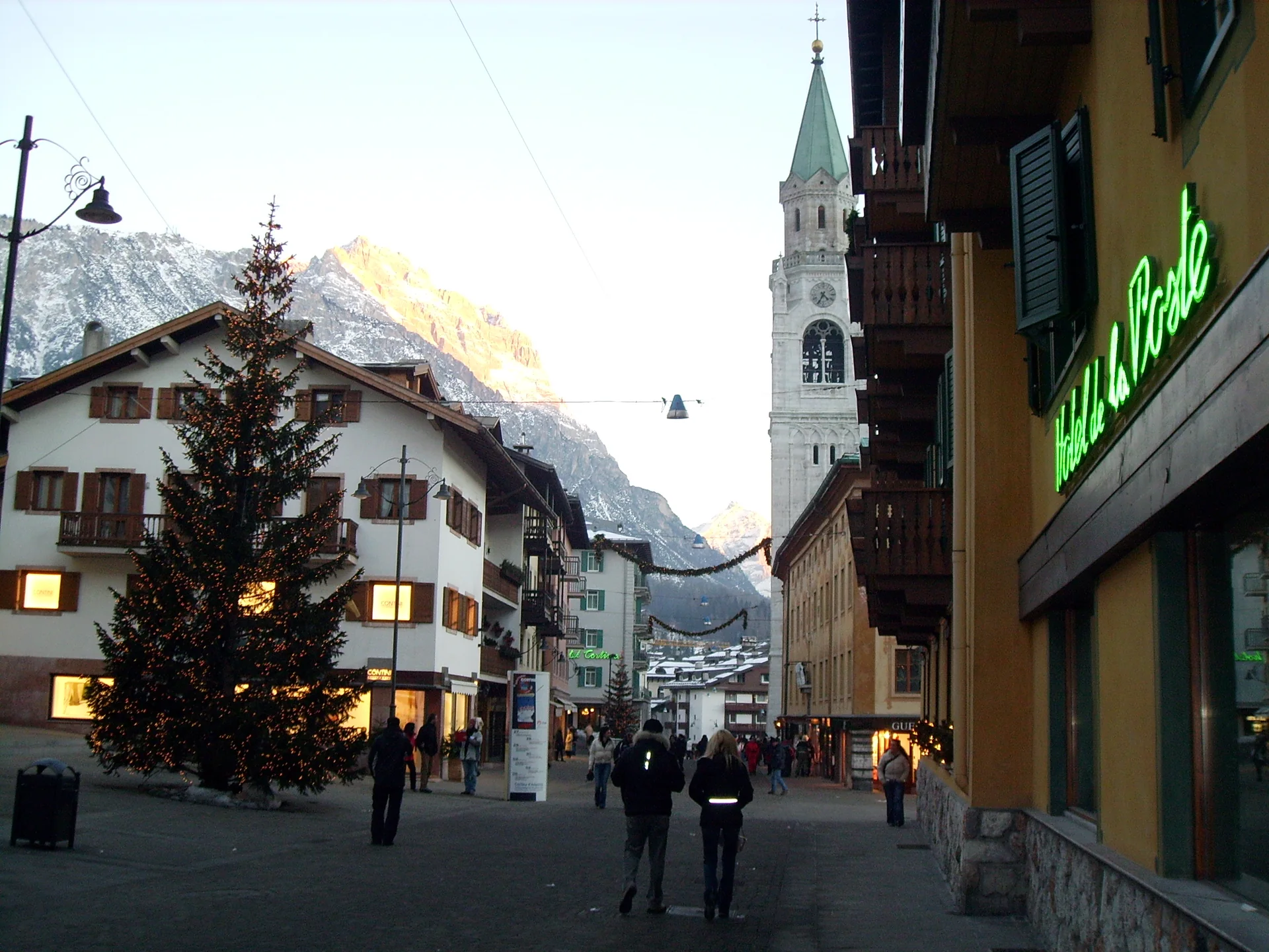 Corso Italia em Cortina d’Ampezzo, com pedestres, torre da igreja e montanhas ao fundo em luz natural.