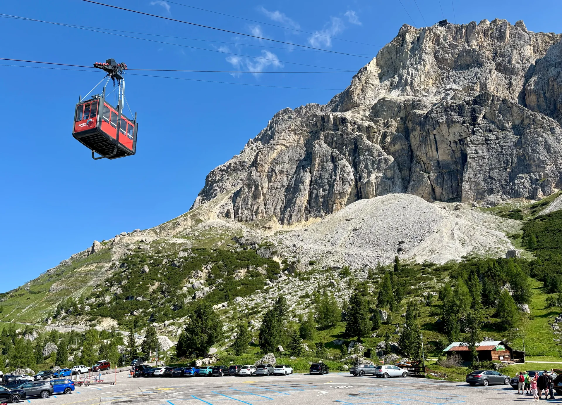 Cabine vermelha do teleférico de Lagazuoi subindo a partir do Passo Falzarego, com paredão rochoso sob céu azul.