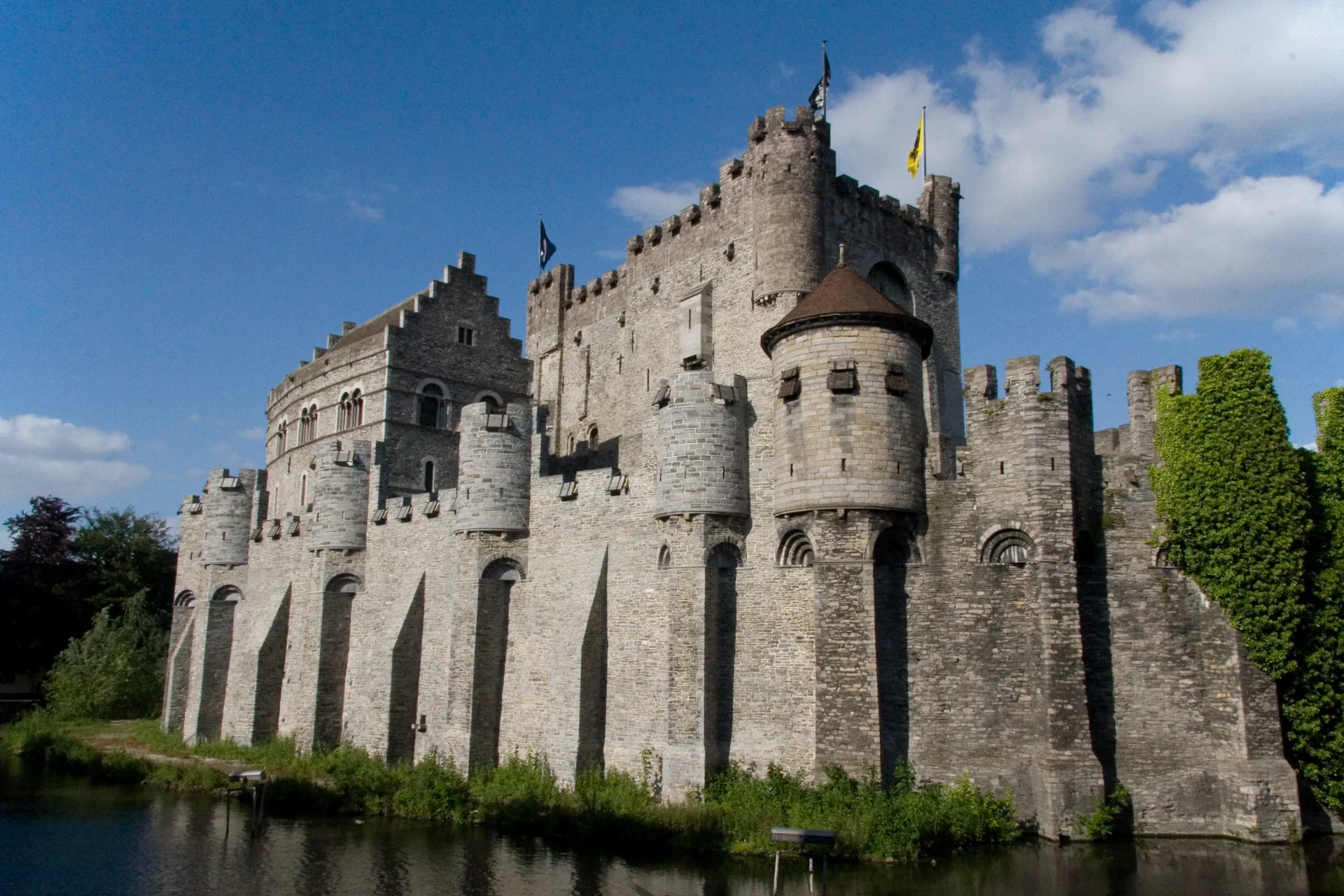 Muralhas, torres e fosso do Castelo Gravensteen em Gante, fotografados sob céu azul.