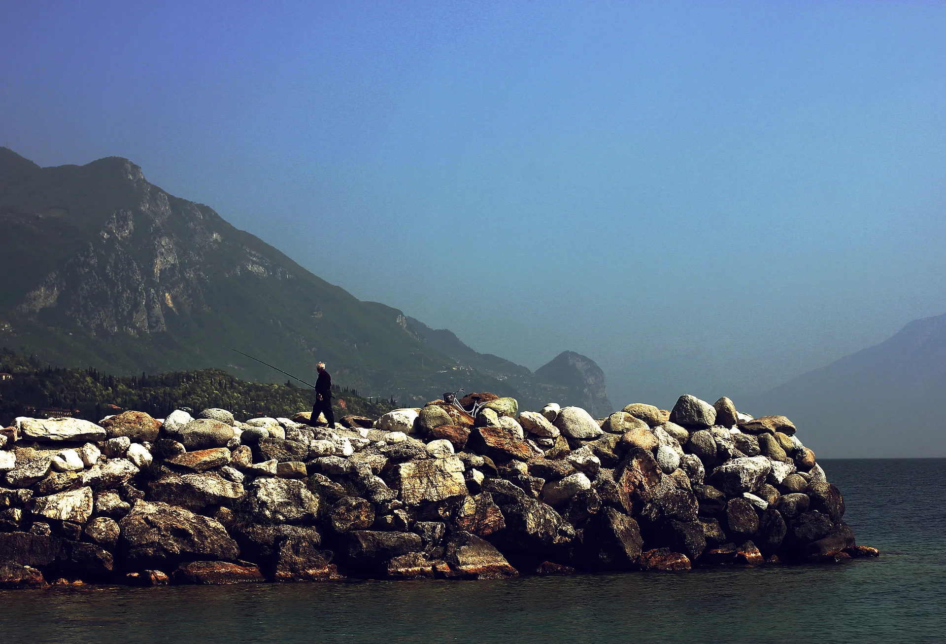 Pescador visto de longe sobre quebra-mar rochoso no Lago di Garda, diante de montanhas e água calma.