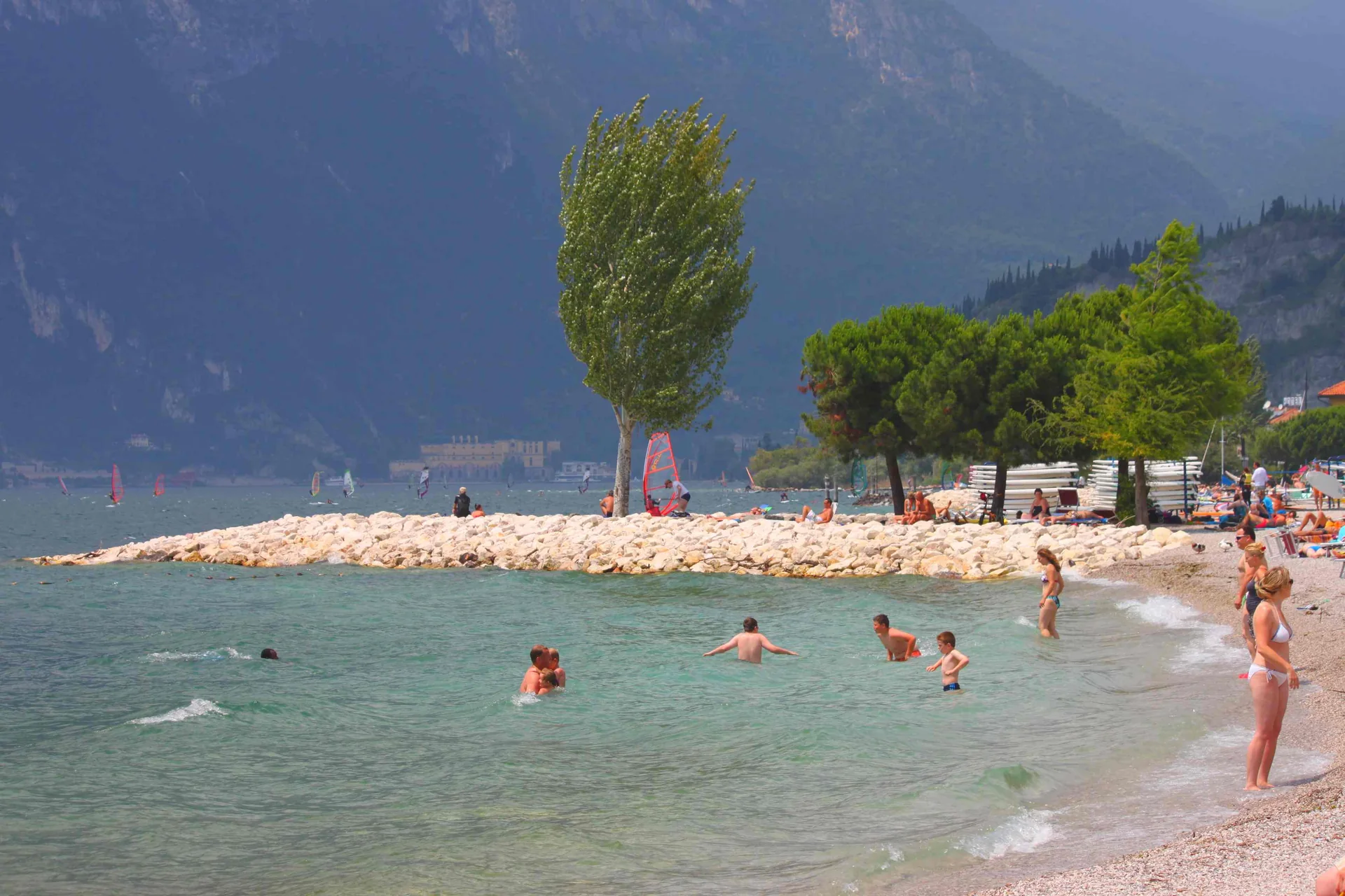 Praia pública de seixos em Torbole, no Lago di Garda, com água e montanhas ao fundo.