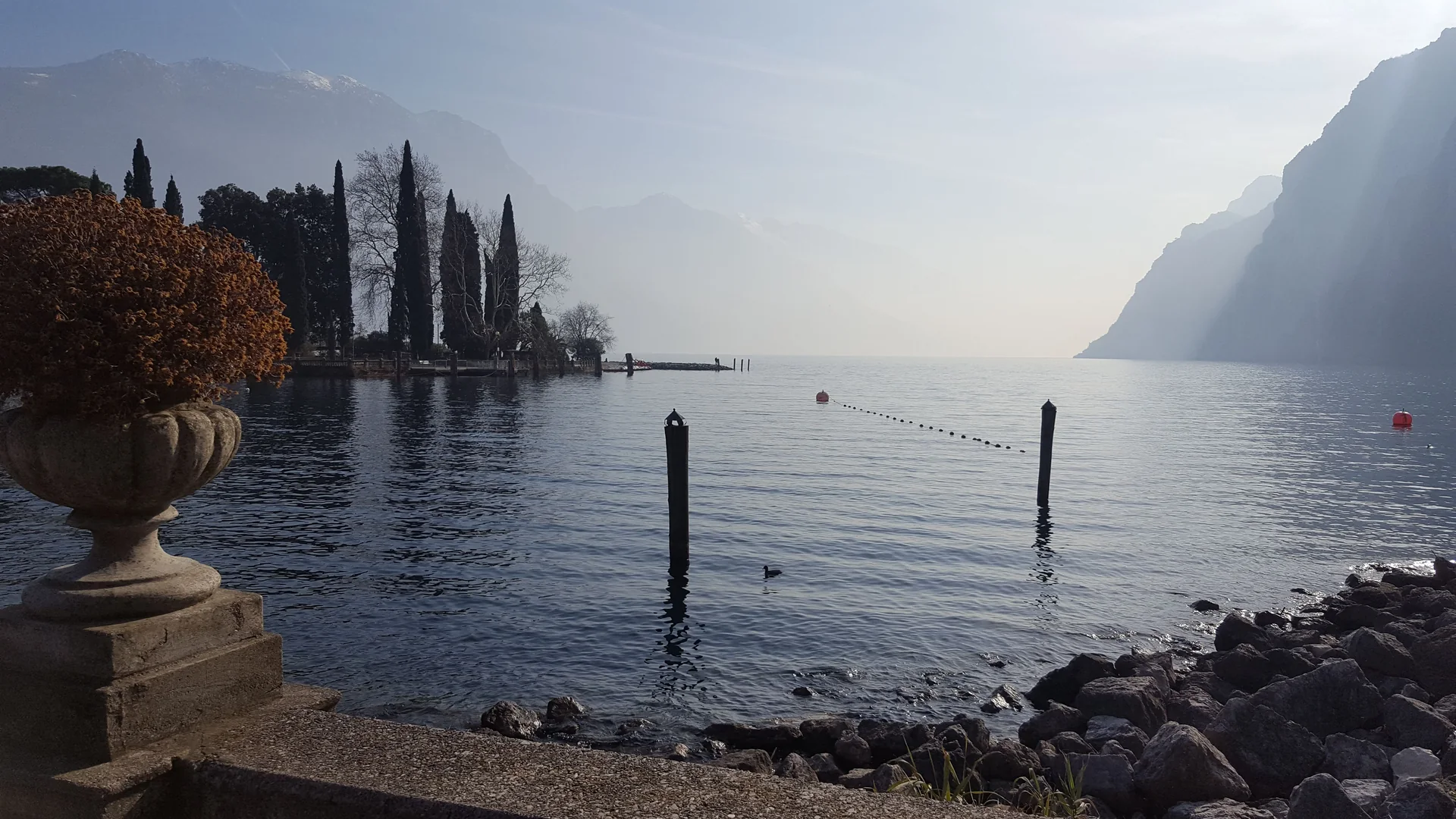 Vista do Lago di Garda no parque Miralago, em Riva del Garda, com margem arborizada e montanhas.