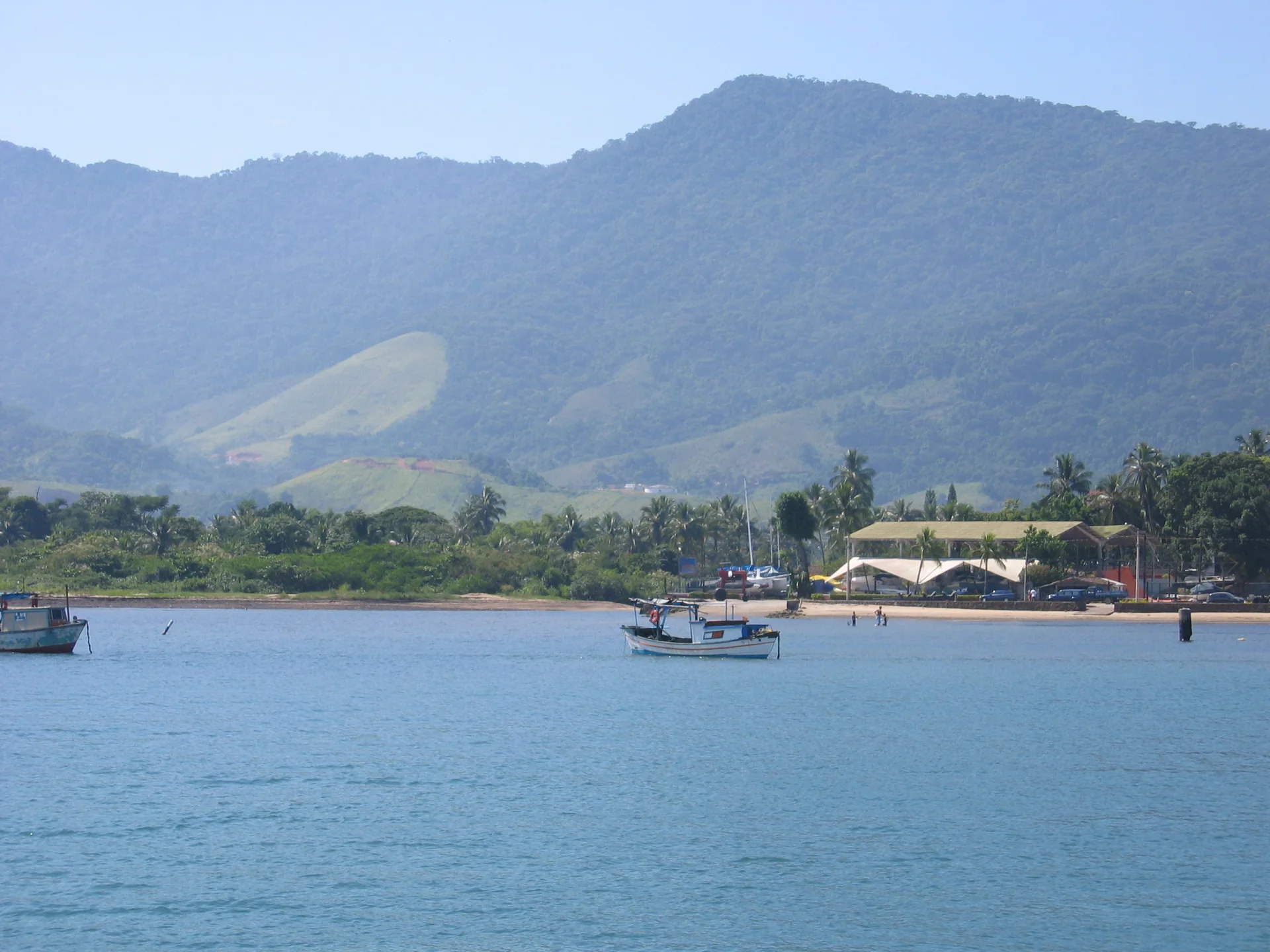 Vista diurna da Praia do Perequê em Ilhabela, observada da balsa que se aproxima da ilha, com mar e encosta verde ao fundo.