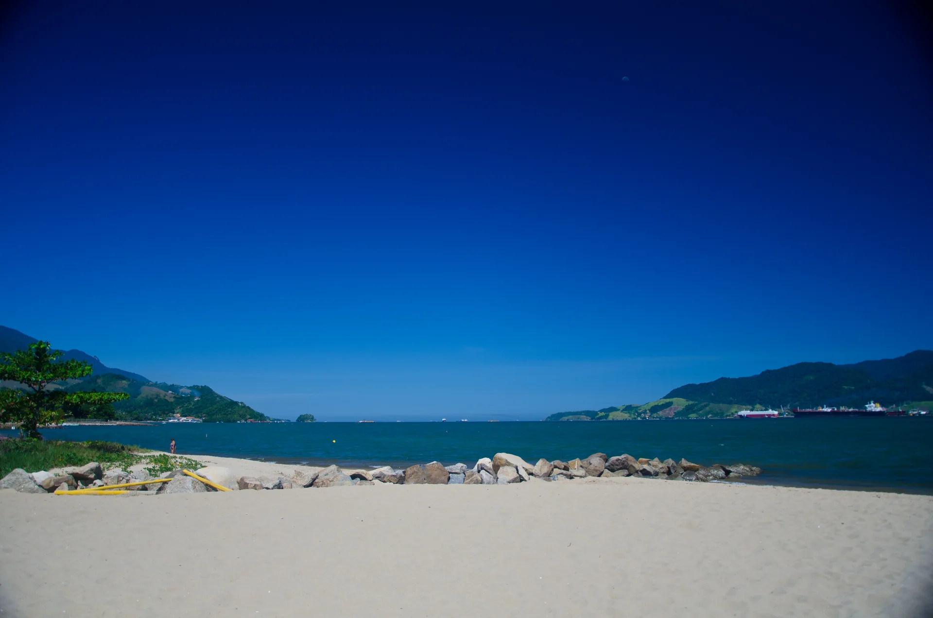 Vista diurna da Praia do Perequê em Ilhabela, com mar do canal, faixa de areia, árvores e montanhas ao fundo.
