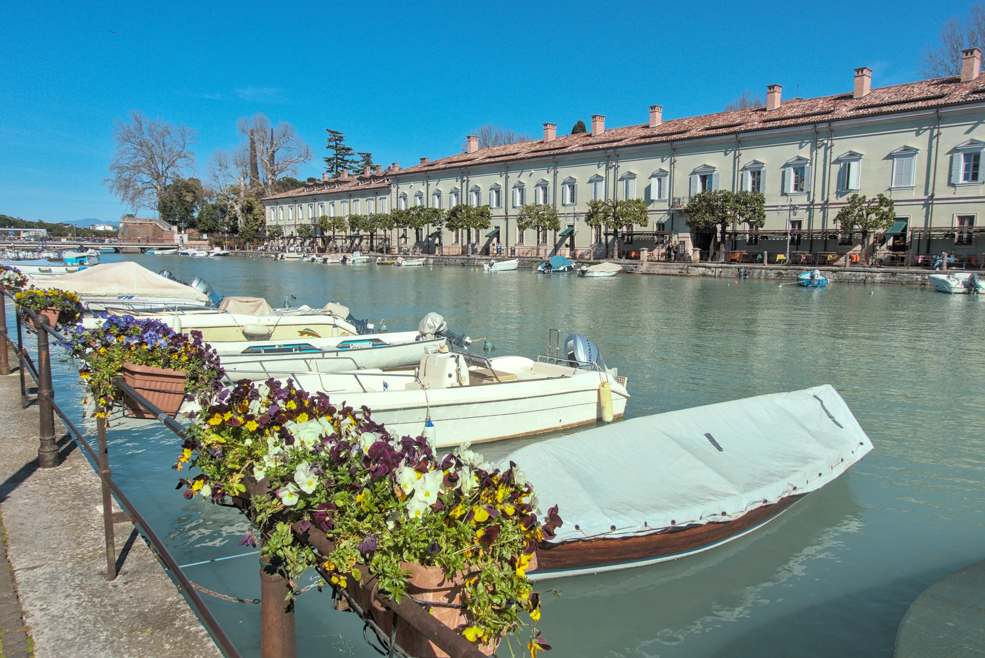 Canale di Mezzo em Peschiera del Garda, com água, barcos e a Caserma XXV Aprile junto às muralhas históricas em dia claro.