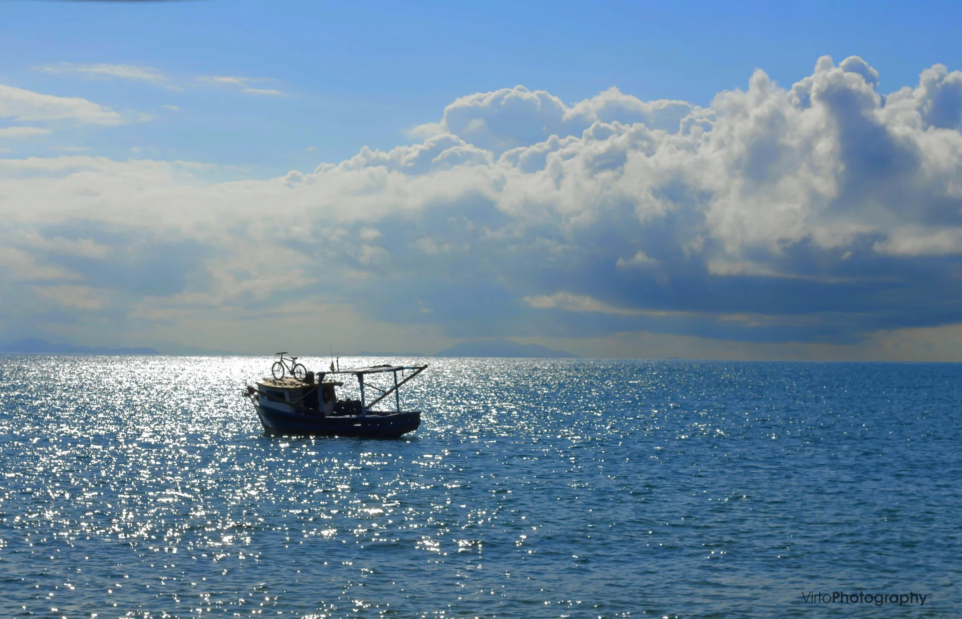 Pequeno barco transporta uma bicicleta no mar azul da Praia do Jabaquara, em Ilhabela, sob céu com nuvens.