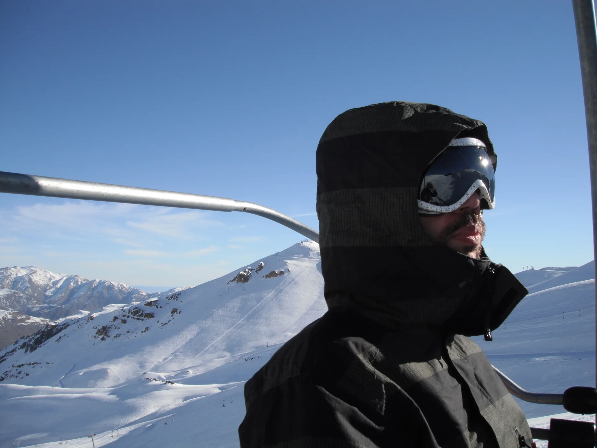Visitante com proteção para o frio e óculos de neve ao lado de um teleférico, com pista nevada e Andes ao fundo.