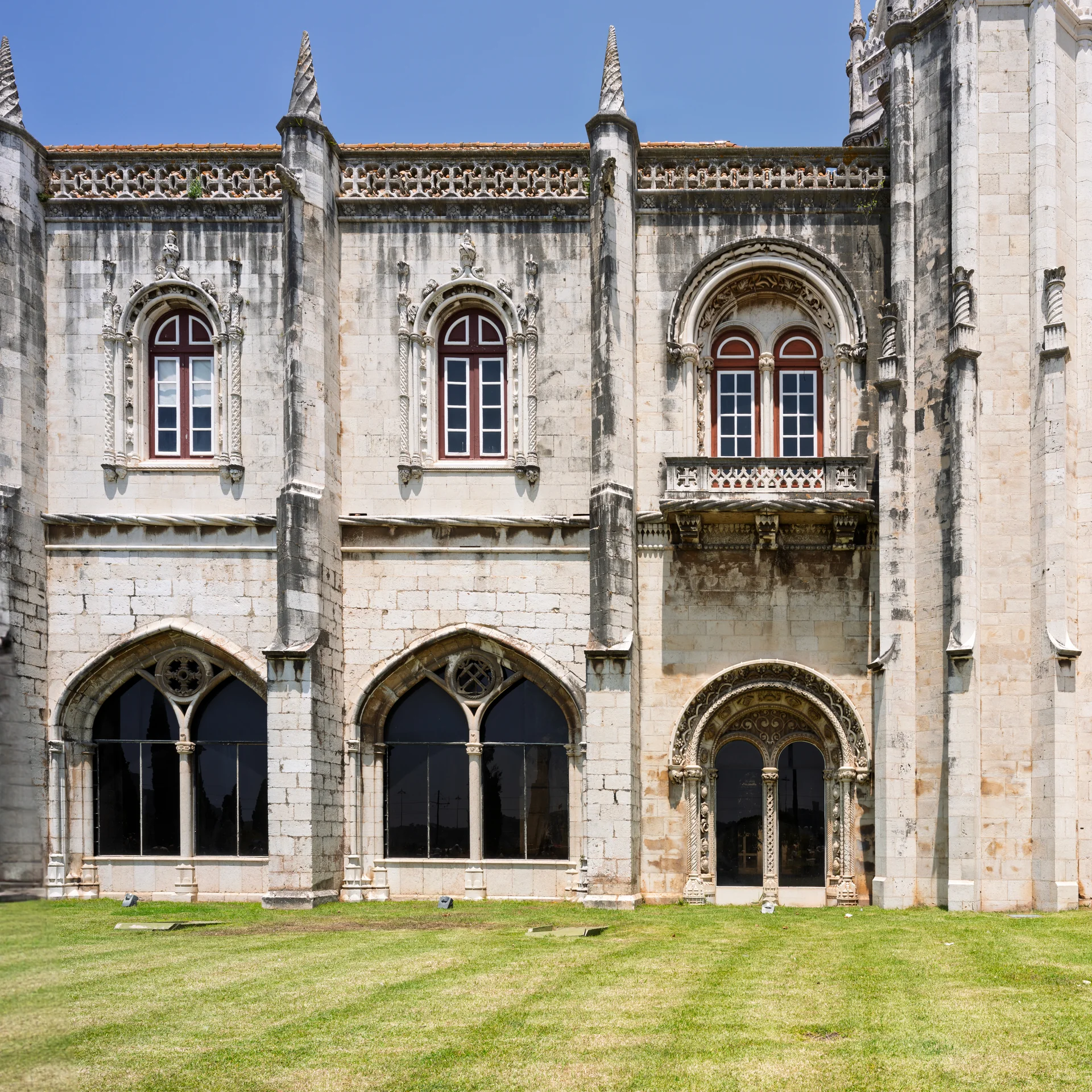 Detalhe de fachada do Mosteiro dos Jerónimos com janelas em arco, cantaria esculpida e pináculos sob céu azul.