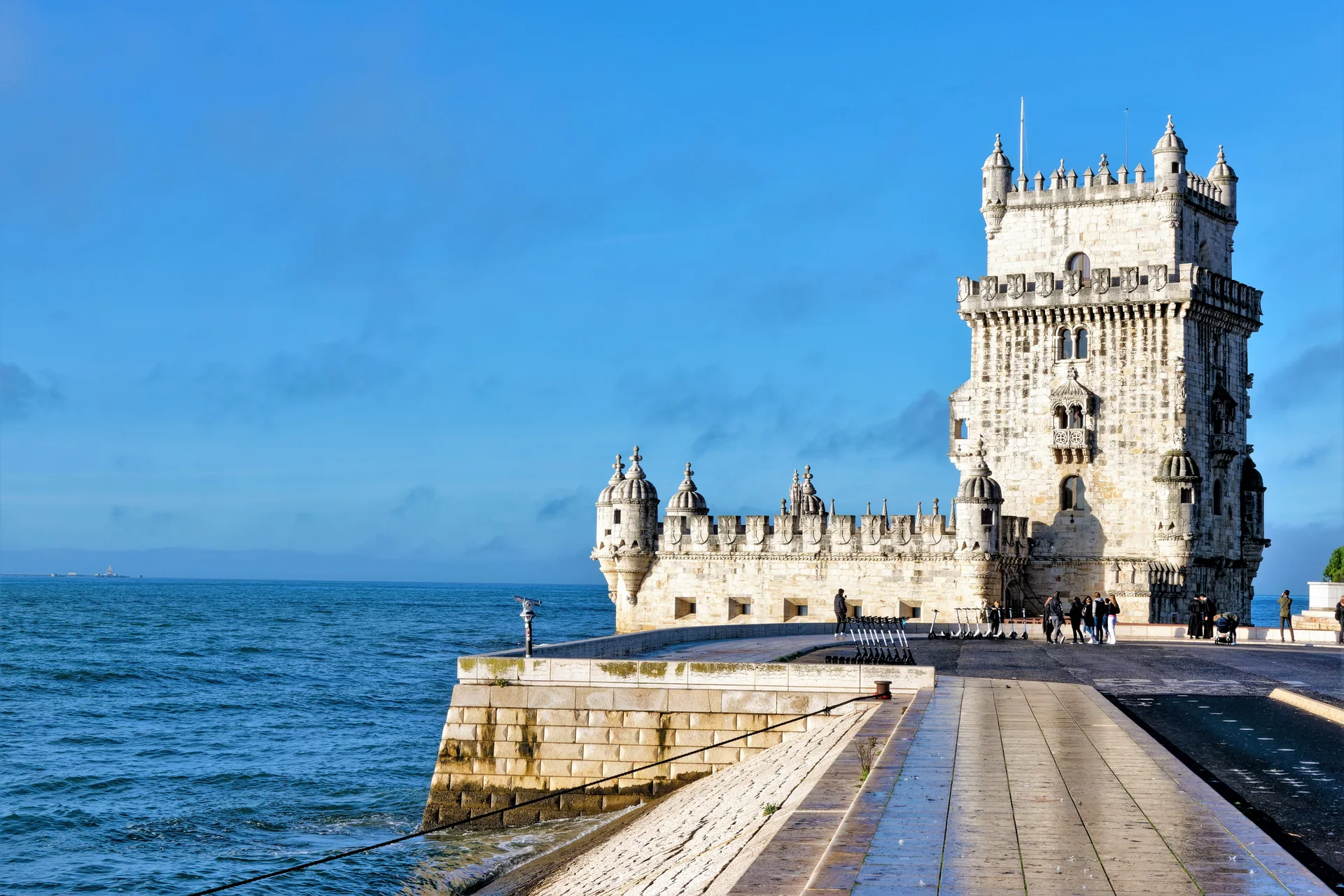 Torre de Belém vista do passeio ribeirinho, com margem do Tejo, cantaria clara e céu azul em Lisboa.