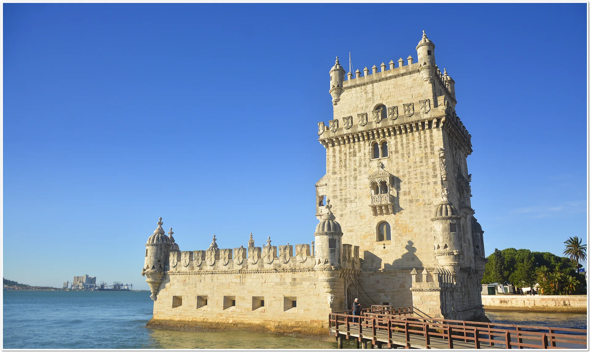 Torre de Belém iluminada pelo sol, com torreão, baluarte de pedra, passarela e águas do Tejo em Lisboa.