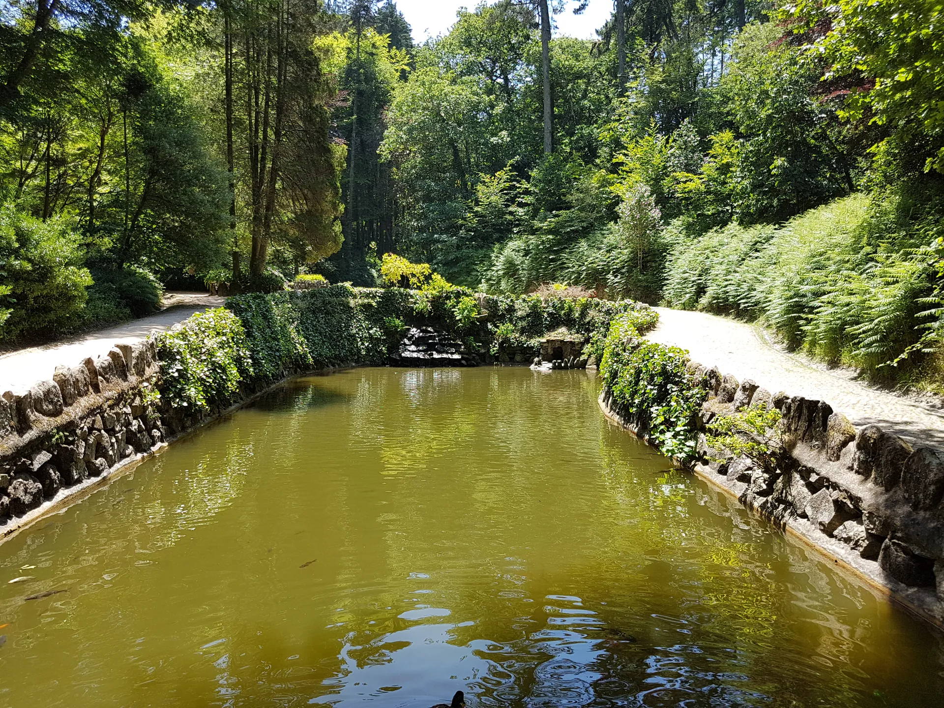 Lago rodeado por muros de pedra, caminhos e vegetação no Vale dos Lagos do Parque da Pena.