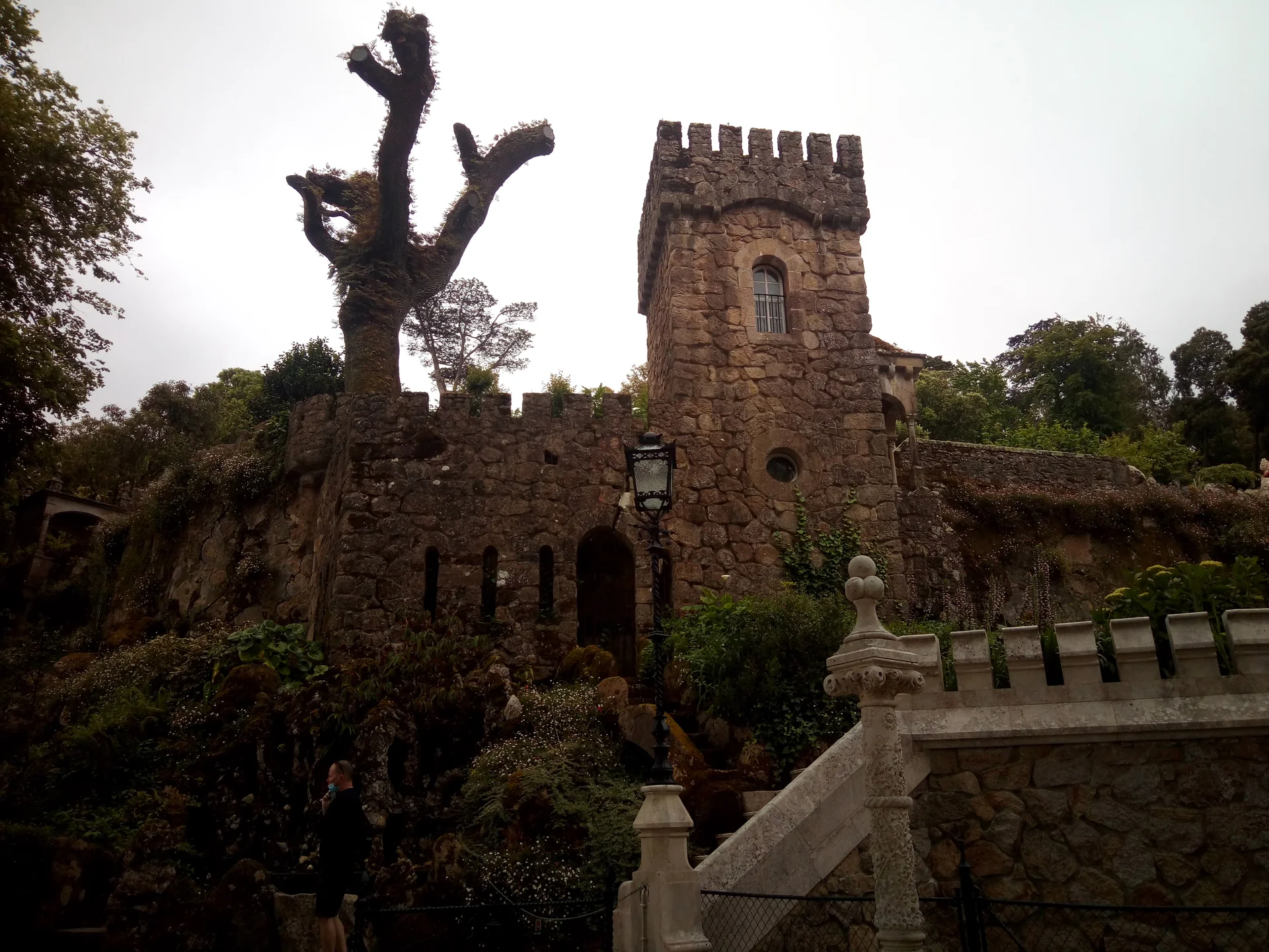 Estrutura de entrada em pedra, torre e caminhos da Quinta da Regaleira entre árvores em Sintra.