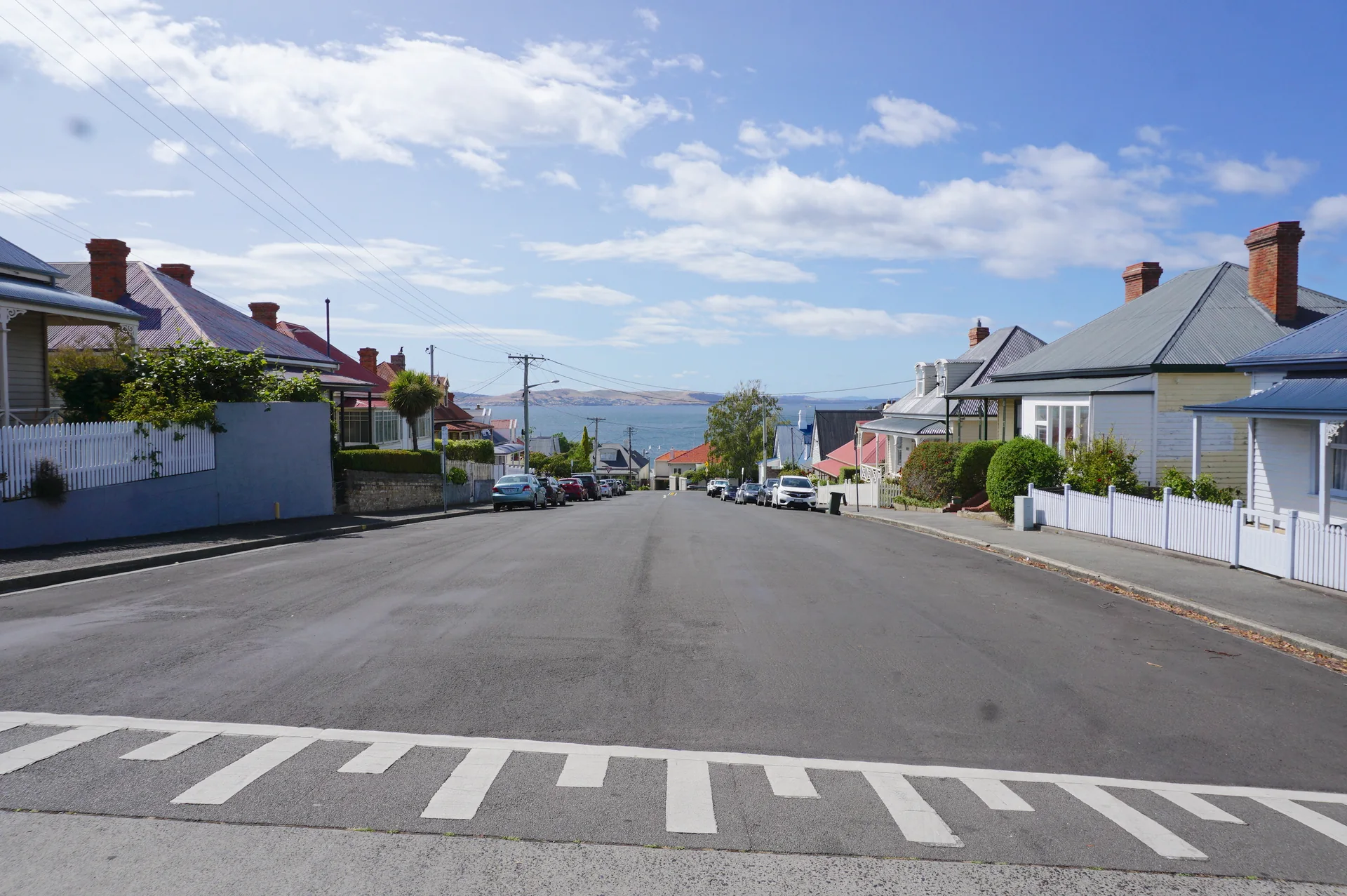 Rua residencial em Battery Point, Hobart, com casas baixas, calçada, carros estacionados e céu claro.