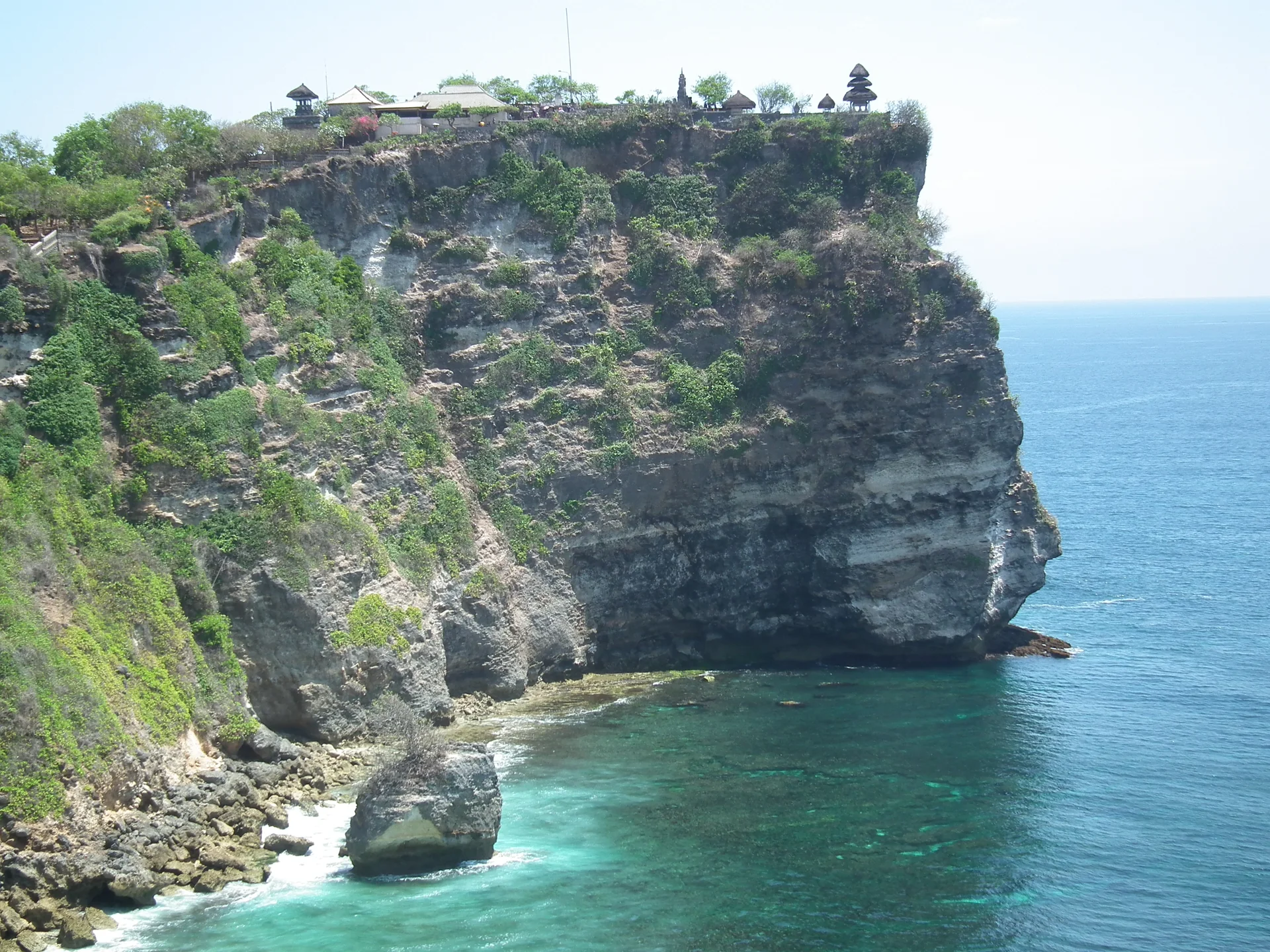 Pura Luhur Uluwatu no alto de uma falésia, com vegetação, paredão rochoso e oceano ao fundo.