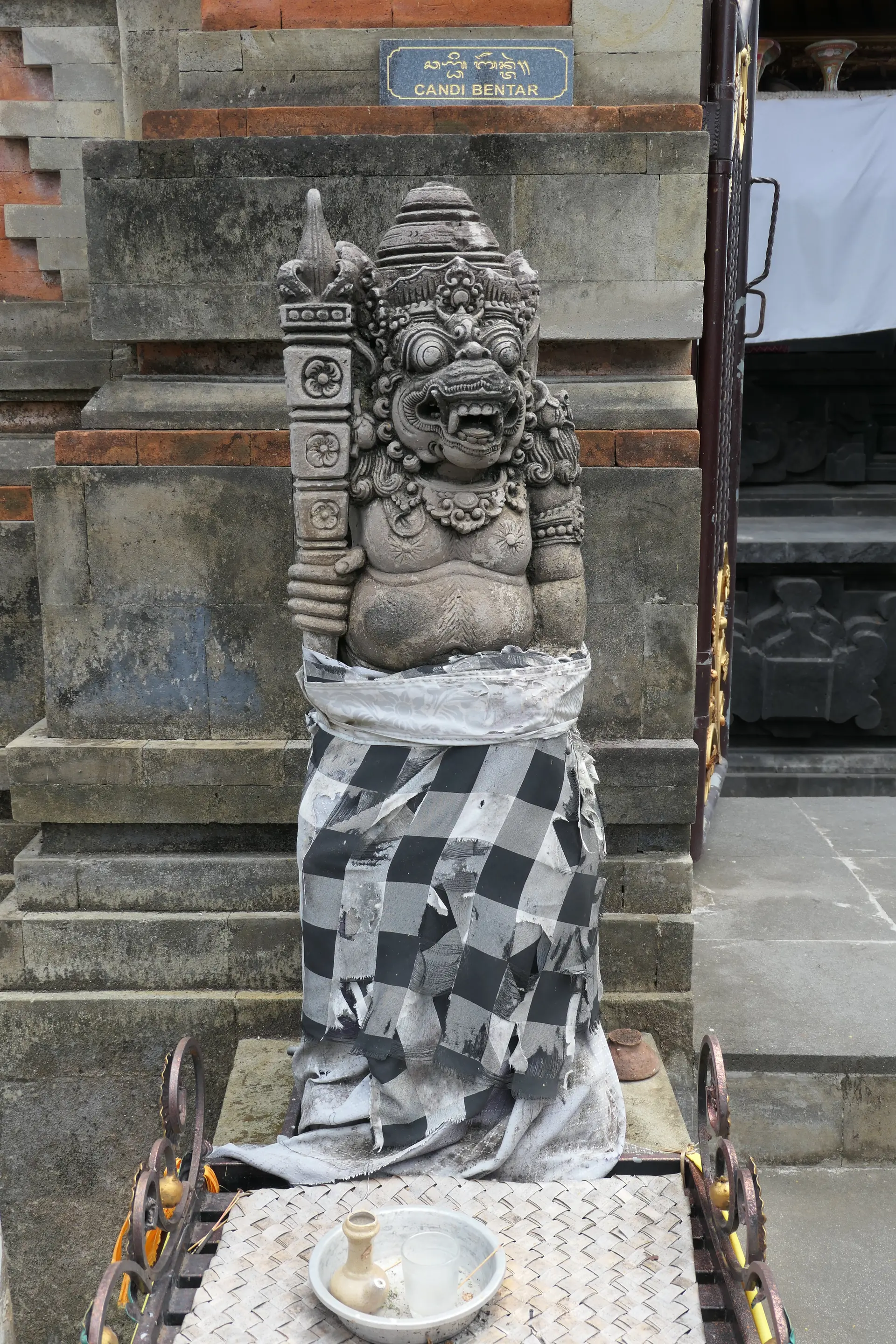 Escultura guardiã junto a um portão de templo em Gianyar, envolta em tecido xadrez e faixa clara, sem pessoas identificáveis.