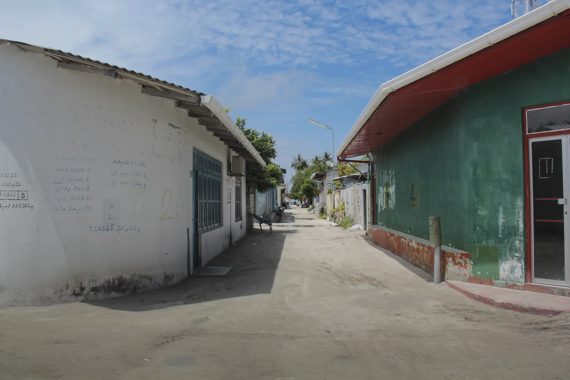 Rua sem pedestres identificáveis em Maafushi, com edifícios baixos, sombra e céu claro.