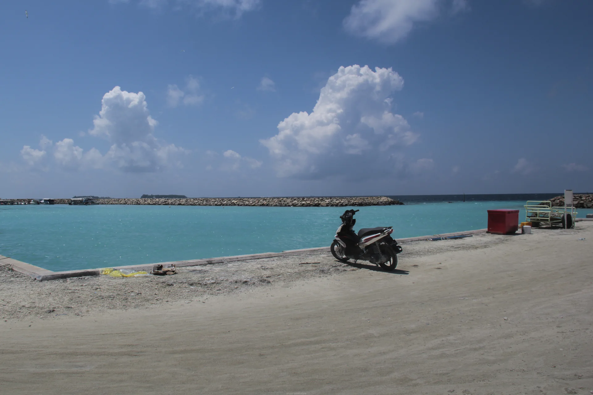 Cais de Maafushi com água azul, molhe baixo e uma motocicleta estacionada, sem trabalhadores ou viajantes identificáveis.
